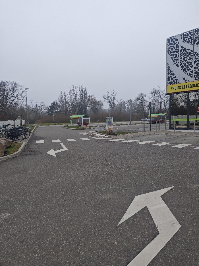 This image captures an outdoor commercial parking area in Hœnheim, France, under an overcast and hazy sky, suggesting low visibility. The foreground features dark grey asphalt pavement with a prominent white L-shaped directional arrow painted on its surface, indicating a left and forward turn. To the immediate right of this arrow, a white dashed-line crosswalk is visible, leading to a textured pedestrian path. On the far left, a grassy median, bordered by large light-colored rocks and a low concrete or stone barrier, accommodates several black bicycles secured together.
In the midground, the asphalt extends into a broader expanse, likely a parking lot. A tall, dark street light post is positioned on the left. Two identical green-roofed structures, appearing to be self-service bays, are situated in the background; one is towards the center-left, and the other towards the center-right. Between these structures, a freestanding sign displays an image of a human figure. A dense line of bare trees forms the distant background, with their branches devoid of foliage.
On the far right, a large building with a distinctive grey and black geometric patterned facade is partially visible. Attached to this building is a bright yellow sign displaying the text "FRUITS ET LÉGUMES" in black capital letters. Multiple vertical metal bollards are installed in front of the building, possibly marking an entrance or pedestrian zone. A dark-colored vehicle is partially discernible in the background, positioned near the rightmost green-roofed self-service bay. No individuals are present, and no discernible actions or interactions are occurring within the scene.