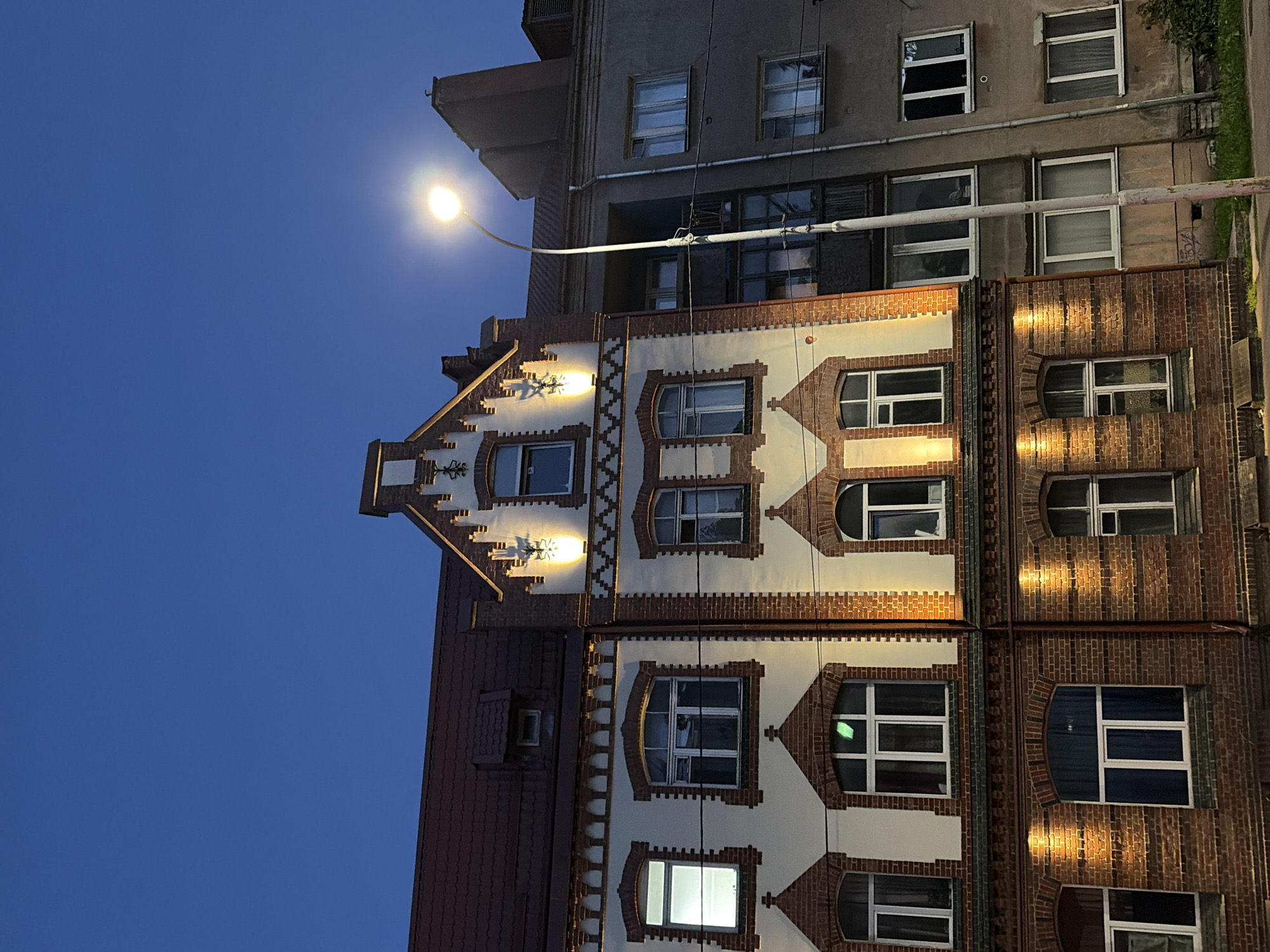 The image shows a multi-story building in a city at dusk. The building has a facade of red brick and white stucco, with decorative brickwork above the windows. There are multiple windows on each floor, some of which are illuminated from within. A street lamp is visible in the foreground, casting a glow on the scene. The sky is a deep blue, indicating the transition from day to night. The building appears to be of historical architecture. The adjacent building is a plain, concrete structure.
