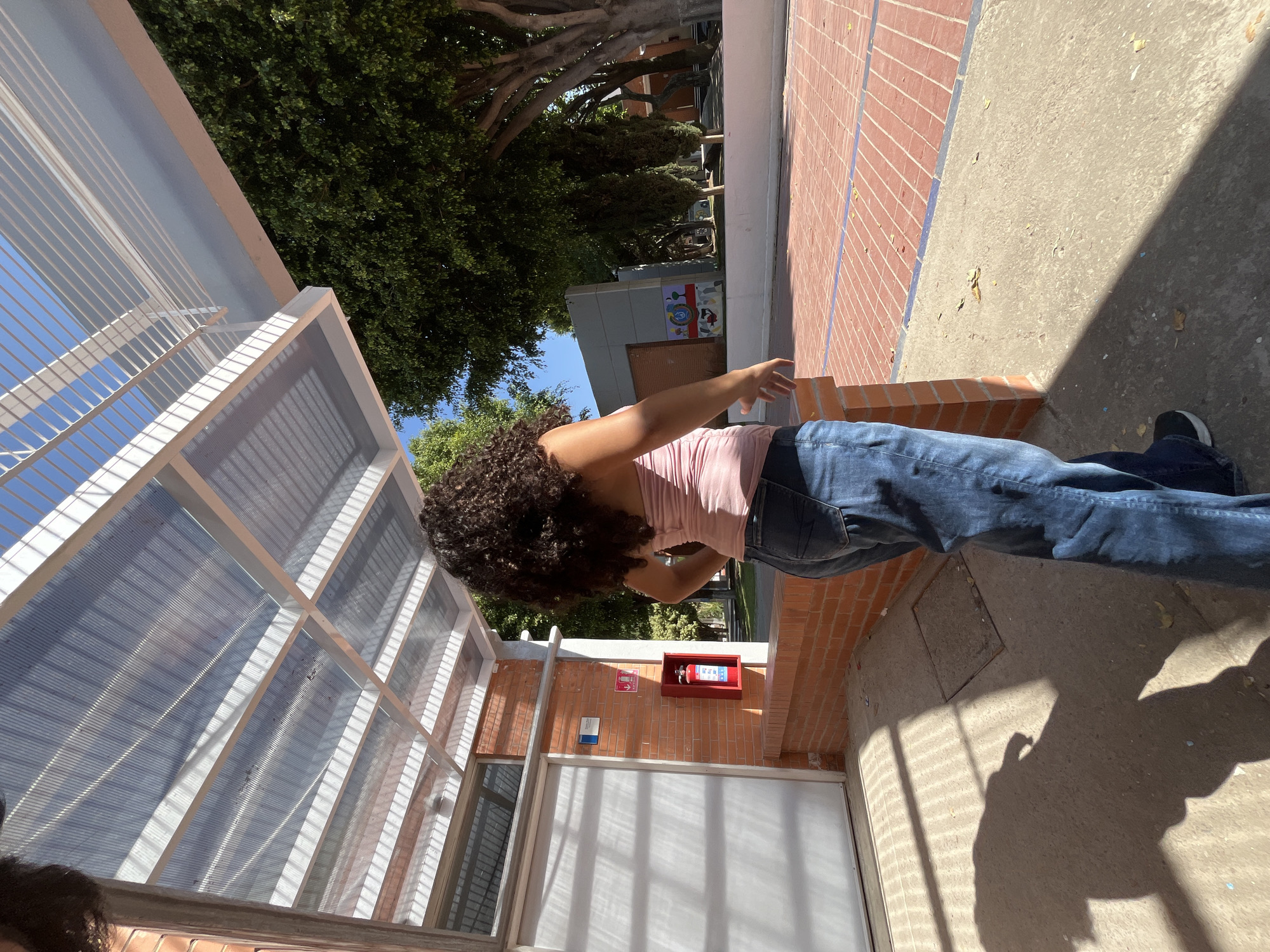 An outdoor scene in Puebla, Mexico, features a person observed from behind, standing under a translucent corrugated plastic canopy supported by a white metal frame. The individual has dark, curly hair and wears a light pink strapless top with blue jeans and black shoes. Their shadow is cast prominently on the concrete ground to the left.
To the immediate left of the person, a brick wall is visible, housing a white roll-up door or panel. Above the door is a small horizontal window. A red fire extinguisher in a wall-mounted box is affixed to the brick wall, beneath a small blue and white sign.
The ground transitions from concrete to a reddish-brown brick-patterned paving with a blue line marking its edge. In the background, numerous large trees with dense green foliage are visible under a clear blue sky. A multi-toned gray building with abstract artwork or graffiti is situated behind the trees and a low white wall, suggesting a campus or institutional environment. Bright sunlight casts distinct shadows across the scene.