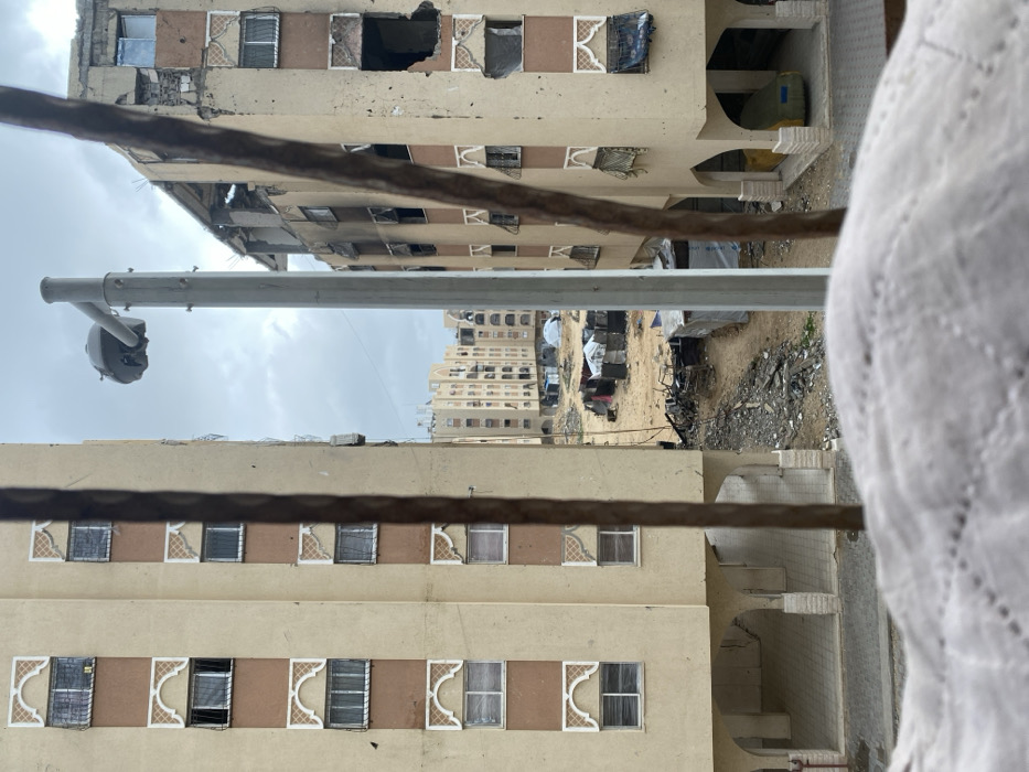 An angled view reveals damaged buildings, one with gaping holes, alongside structures with intact windows. A light pole is visible against a cloudy sky. Debris-strewn ground contrasts with tent-like shelters nearby. A patterned white fabric occupies the right foreground, partially obscuring the urban landscape. The composition highlights a stark contrast between destruction and resilient human presence.