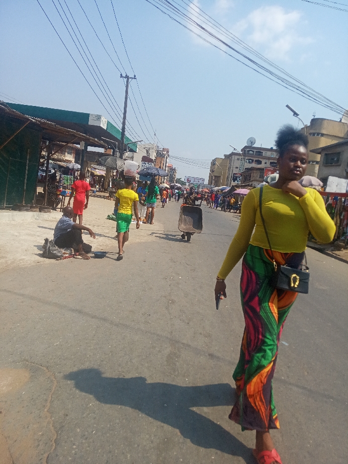 The image depicts a bustling daytime street scene in Nkwelle Ezunaka, Nigeria. The paved asphalt road shows signs of wear, with visible cracks and variations in texture. Strong overhead sunlight casts distinct shadows across the pavement.
In the foreground, slightly to the right of center, a female subject walks towards the viewer. She wears a long-sleeved, sheer yellow top, a long skirt with a vibrant multi-colored pattern (green, red, orange, black), and red open-toed sandals. A small black shoulder bag with a gold horseshoe-shaped clasp hangs from her left shoulder. She holds a dark-colored smartphone in her right hand, while her left hand is raised towards her neck area. Her dark, curly hair is styled upwards.
In the midground, to the left, a barefoot man sits directly on the pavement, dressed in a light blue short-sleeved collared shirt and dark trousers. A small dark bag or bundle rests beside him, and a small blue bowl is on the ground in front. Further down the street, multiple pedestrians are visible, moving away from the viewer. These include an individual in a bright red dress, and another in a yellow t-shirt and green shorts carrying a white container on their head. A dark-colored wheelbarrow is also present, appearing to be pushed or pulled.
The background on the left features a low-lying structure with a green and white striped canopy, possibly part of a gas station or a large market stall. Numerous roadside stalls or small shops line the left side of the street, many with corrugated iron roofs and displaying merchandise. On the right side of the background, multi-story buildings with various architectural details are visible, some featuring satellite dishes. A utility pole supporting multiple overhead power lines stands prominently in the mid-left. The sky above is clear and blue with minimal cloud cover.
