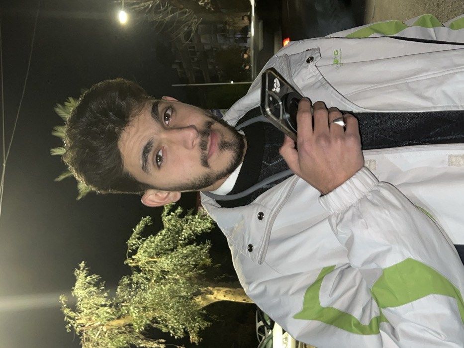 A young man with dark hair and a beard poses outdoors at night. He holds a smartphone to his ear with his right hand, where he wears a silver ring. He is dressed in a white jacket with bright green accents, layered over a dark sweater and a collared shirt. Behind him are dimly lit trees and a building in the background. The scene is lit with soft, ambient lighting.