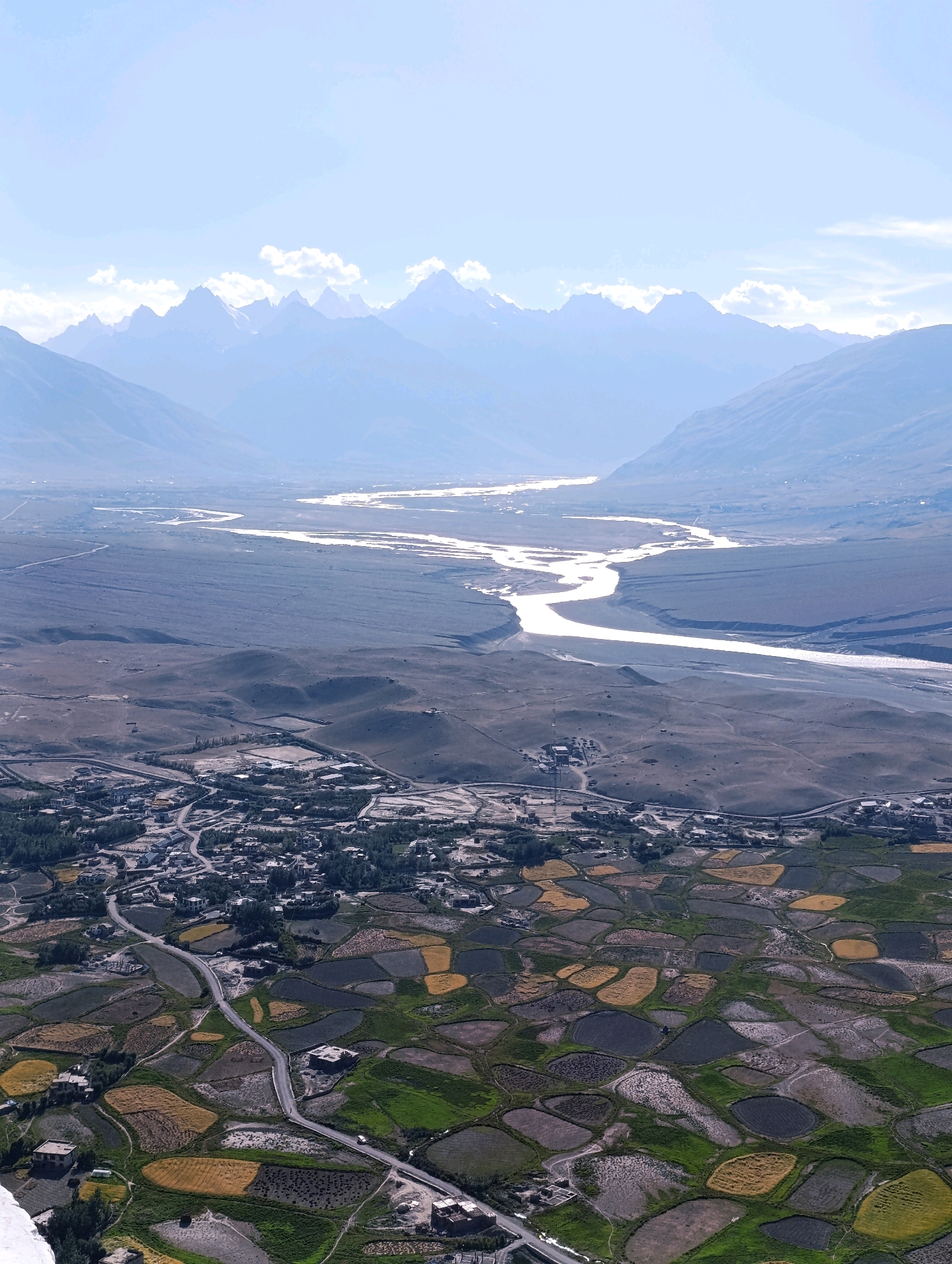 The image depicts a wide aerial view of the city of Pibiting, India, with a sprawling landscape that includes agricultural fields, a town, a river, and mountains in the distance. The foreground is dominated by patchwork fields of varying colors, suggesting different crops or stages of growth. These fields are irregularly shaped and are interspersed with dirt paths and some buildings.
A winding road runs through the agricultural area, leading towards a more densely populated town. The town itself appears to be composed of many small buildings with dark roofs, clustered together and connected by a network of roads and paths. Some of these buildings have courtyards or open spaces.
Further in the mid-ground, a wide, sandy river snakes through a vast, barren plain. The riverbed is wide, with the water appearing bright and reflecting the sky. The plain surrounding the river is mostly muted in color, with some undulations and scattered structures that appear to be industrial or rural outbuildings.
In the background, a majestic range of mountains rises, their peaks softened by distance and a light haze. The mountains are layered, with the farthest ranges appearing almost ethereal against a pale blue sky with scattered clouds. The overall impression is one of a vast, arid landscape brought to life by human settlement and agriculture, set against a dramatic mountainous backdrop. No people are directly visible in the image.