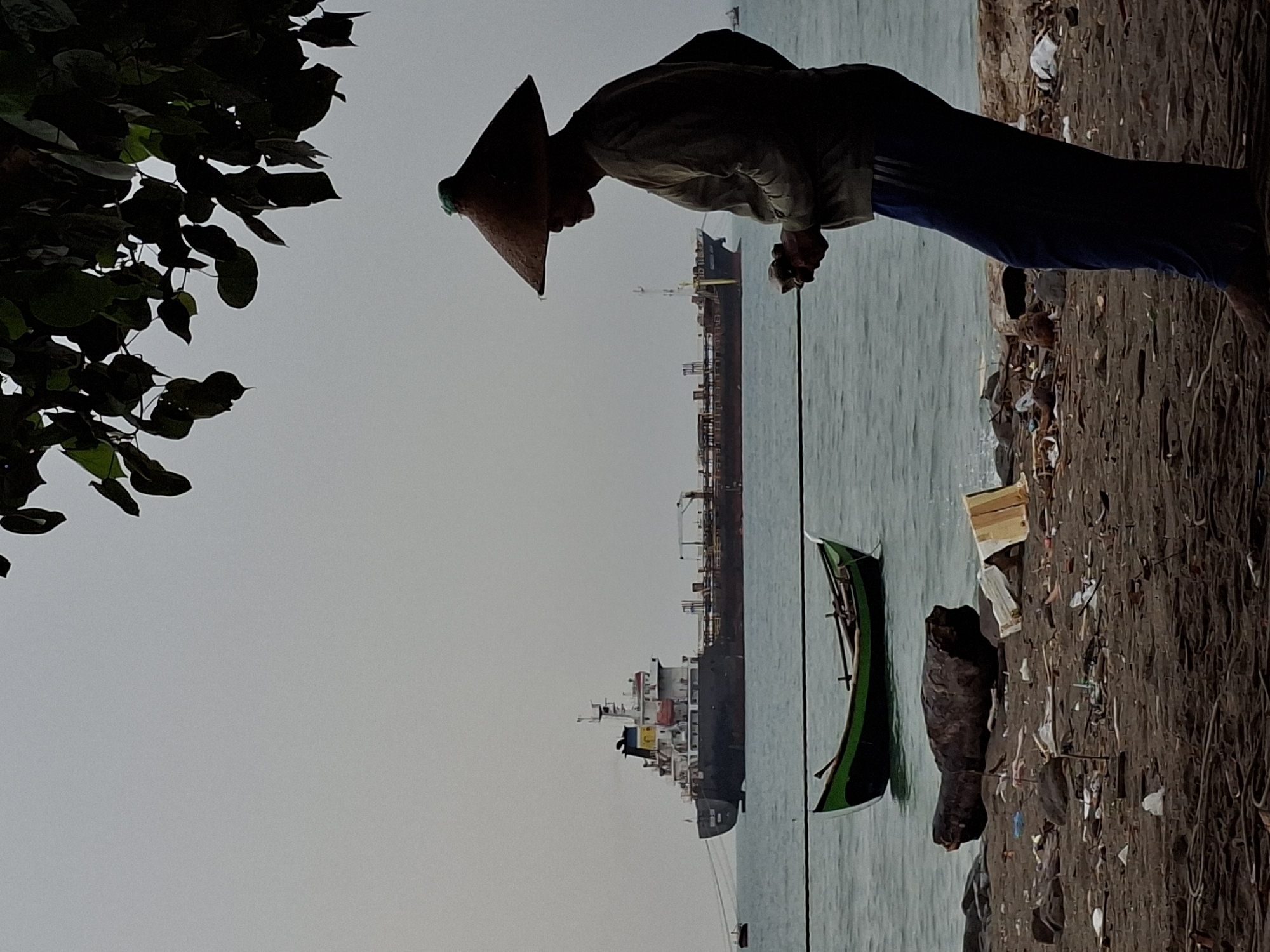 A silhouetted figure wearing a conical hat stands on a debris-strewn beach in Lhokseumawe, Indonesia. The figure, seen from the side, is wearing a light-colored long-sleeved shirt and dark blue pants. Their hands are clasped together in front of them. Behind the figure, the sea is visible with two boats. The closest boat is a small, green and black fishing vessel with oars. Further in the background, a large cargo ship is anchored on the water. The sky is overcast. The beach itself is covered in sand, rocks, and various pieces of trash, including plastic bags, wood debris, and cardboard. The overall scene suggests a coastal environment, possibly a working harbor or a fishing area, marked by both marine activity and the presence of litter.
