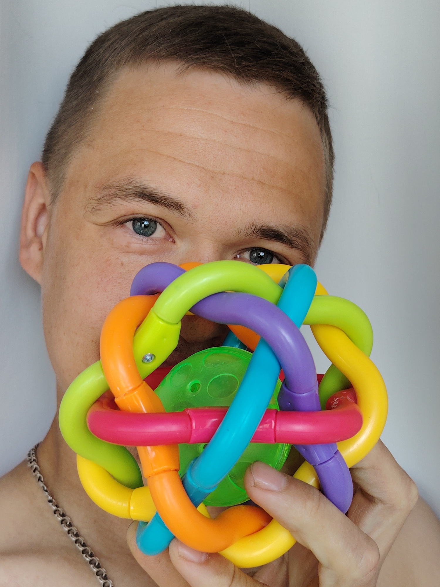 A person with light skin, short brown hair, and blue eyes is holding a colorful, interconnected, spherical toy in front of their face. The toy is made up of many curved, plastic tubes of various colors, including green, orange, yellow, pink, blue, and purple. A green plastic sphere is visible in the center of the toy. The person's eyes are visible above the toy. A silver chain is visible around their neck. The background is a plain white wall. No information suggests that this image was taken in Chaikovsky, Russia.