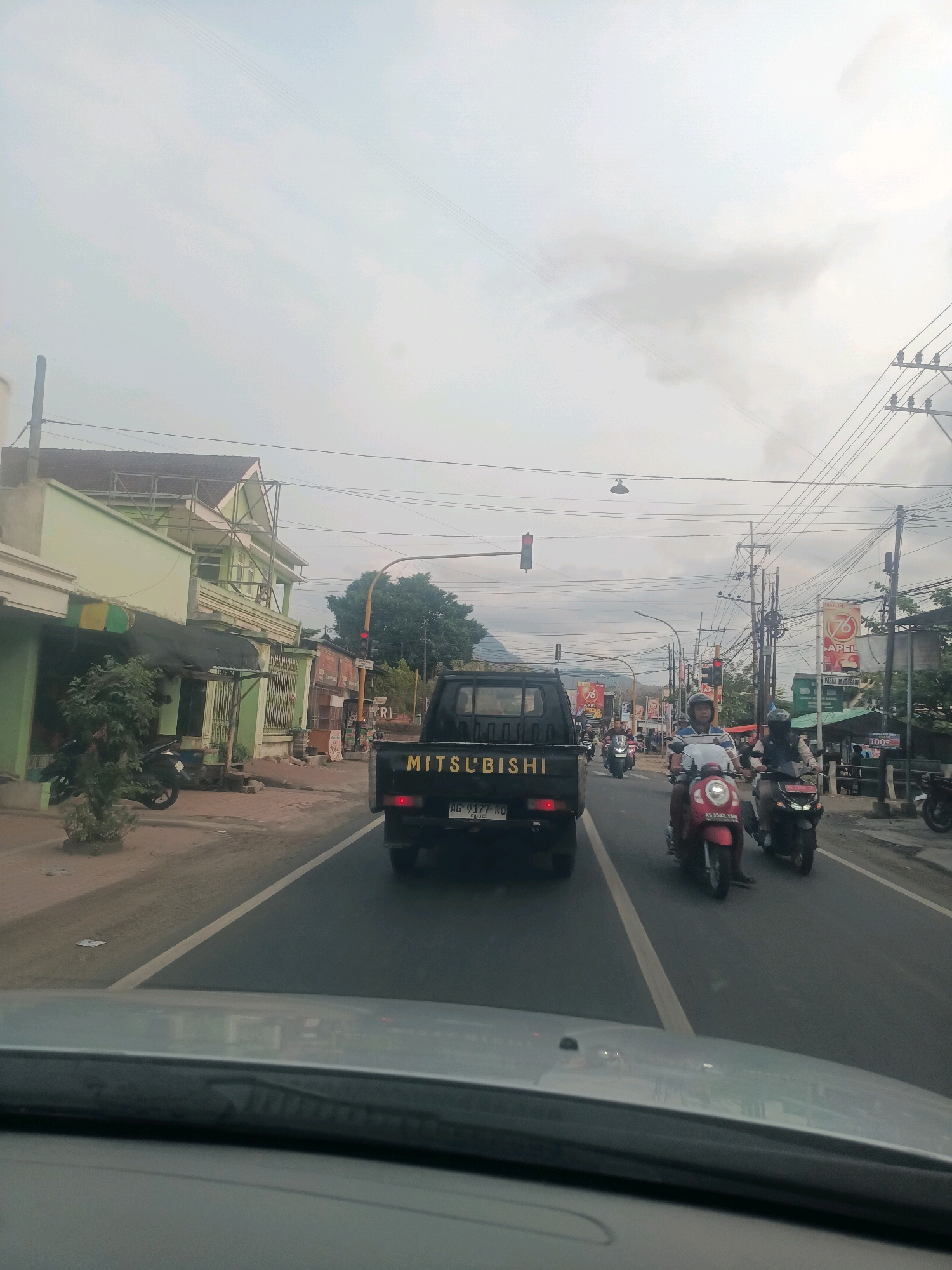 The image shows a street scene in Gandusari, Indonesia, likely during daylight hours, with overcast skies. The perspective is from inside a vehicle, looking forward.
In the foreground, the dashboard and windshield of the vehicle are visible. The road ahead is paved and marked with a yellow line.
Dominating the center of the image is a black Mitsubishi pickup truck with yellow lettering on the tailgate. The truck has a license plate reading "AG 9177 RU". Behind the truck, several motorcycles are traveling in the same direction. One red scooter is particularly prominent, with a rider wearing a helmet. Another motorcycle, dark in color, is to its right, also with a rider wearing a helmet. Further back, more motorcycles are visible in the distance.
To the left of the road, there are buildings with light green facades. One building has an upper floor with balconies and a metal framework. A black motorcycle is parked in front of one of the buildings. Trees and vegetation are also present along the roadside.
To the right of the road, there are more buildings, some appearing to be shops with awnings. Signs are visible on some of these buildings, including one that says "76 APEL". Utility poles with numerous wires are scattered throughout the scene, a common sight in urban or semi-urban Indonesian environments. A traffic light is visible, currently red. The overall impression is of a moderately busy street with typical urban infrastructure.