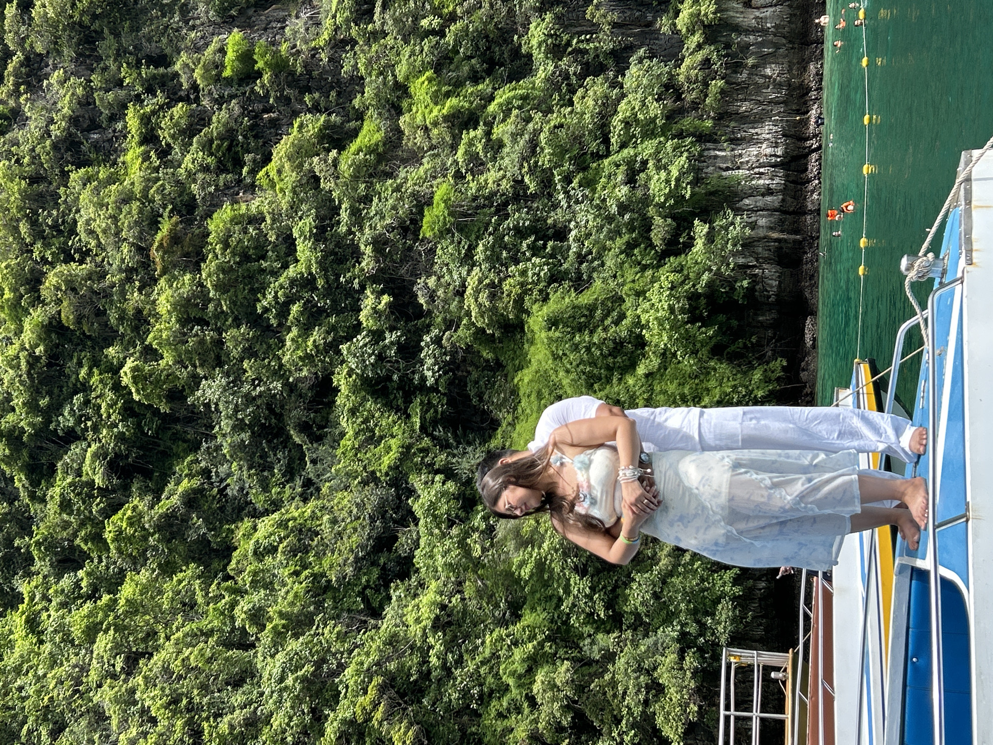 In Ao Nang, Thailand, a couple stands embracing on the deck of a boat. The woman, wearing sunglasses and a floral top with a light blue and white patterned skirt, is positioned in the foreground, her arms wrapped around herself. Behind her, a man in a white outfit stands with his arms around her waist. The boat, with its blue, white, and yellow accents, rests on turquoise water. In the background, a dense, lush green cliffside rises, covered in vegetation and trees. Several other boats are visible, and people can be seen swimming and enjoying the water.