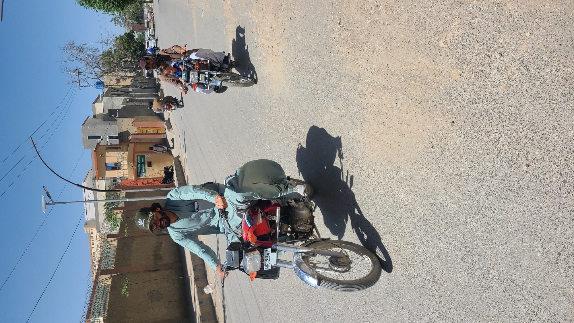 The image depicts a street scene in Quetta City Tehsil, Pakistan. A man wearing a light blue shalwar kameez and an Adidas baseball cap is riding a red motorcycle with the license plate "MR-2347" on a paved road. In the background, another motorcycle with three people, including at least one child, is traveling in the same direction. Buildings are visible on either side of the road. A street light stands near a wall topped with barbed wire. The sky is clear and blue. Shadows suggest it is daytime.