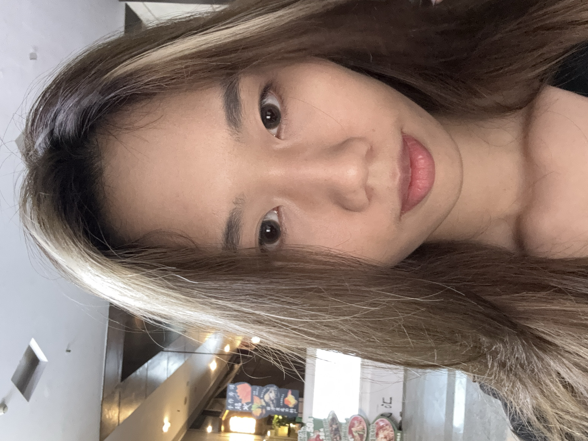 A medium shot photograph depicts a young woman with brown hair and light blonde highlights. She is facing the camera at an angle, with a soft, almost smiling expression. The background includes interior architectural elements, possibly a restaurant or food establishment. In the background, we see signage with food images and Chinese characters, indicating a likely location in China. The lighting is warm, suggesting an indoor setting with artificial lighting. Based on the Chinese characters on the menu, this might be near a food establishment in Yuzhong District, China.