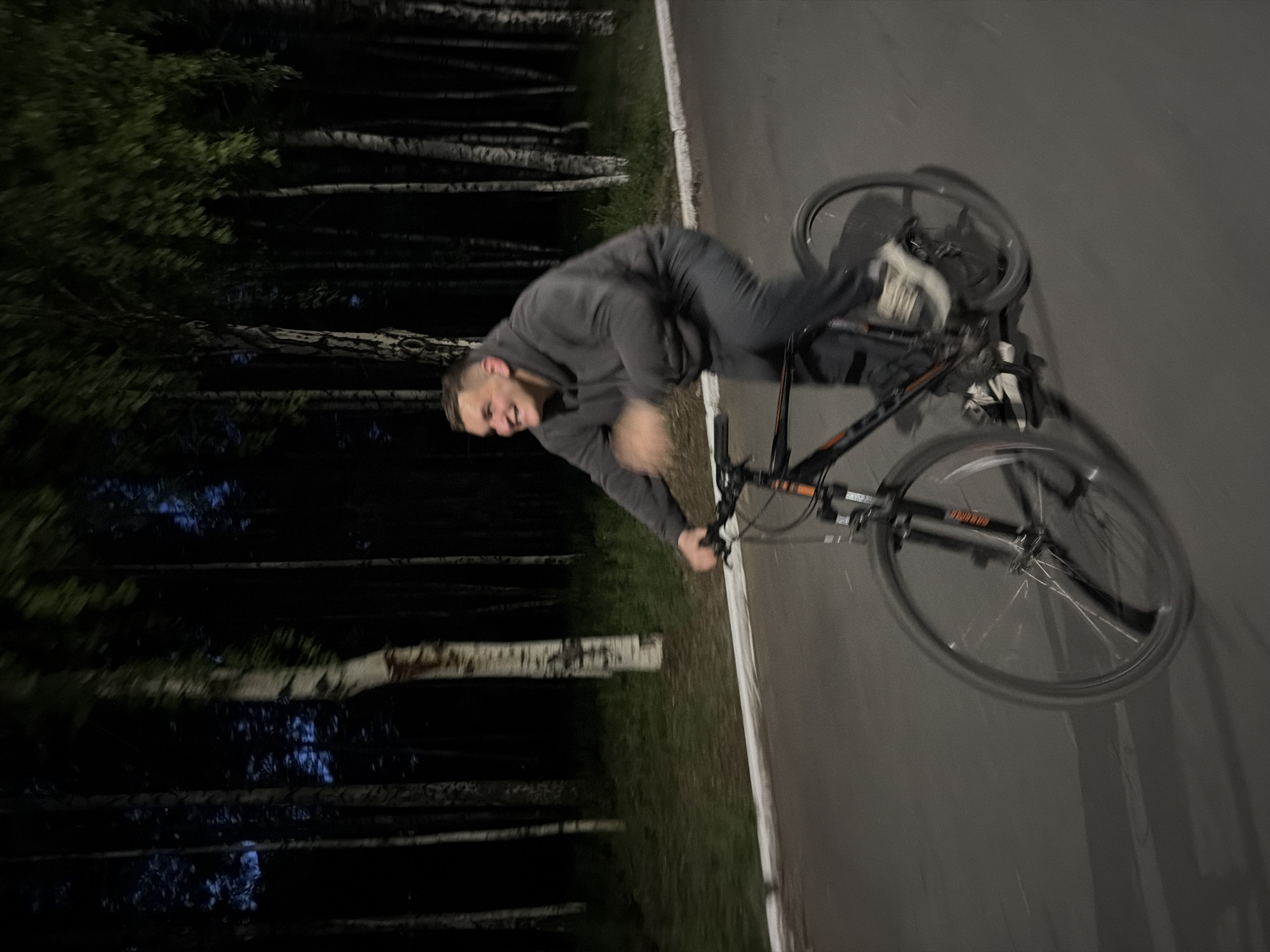 The image displays a dynamic nighttime scene of a young man riding a bicycle on a paved path next to a forest. The man, who appears to be in his late teens or early twenties with short dark hair and a beard, is captured mid-action, leaning significantly as if performing a trick or sharp turn. He is smiling broadly, conveying a sense of enjoyment or concentration. He wears a dark grey hoodie, dark trousers, and white sneakers.
The bicycle has a black frame with some orange detailing. Its wheels are visibly blurred, emphasizing the motion. The paved surface features a white line along its edge. To the side of the path, a dense forest stands in deep shadow, with the distinct white bark of several birch trees visible against the dark background. The dim lighting confirms that the photograph was taken at night or very late in the evening. There is no visible artificial lighting source, contributing to the dark, atmospheric setting. While some text is present on the bicycle frame and tires, it is too indistinct to be read. The scene takes place in Izhevsk, Russia.