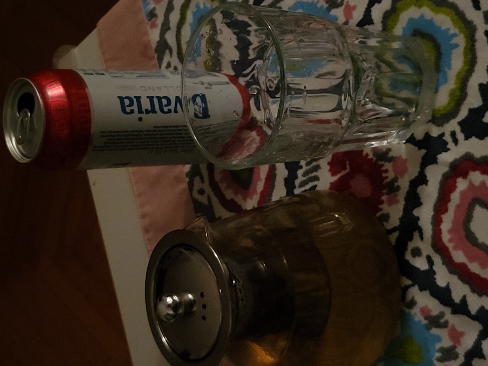 A can of Bavaria beer, an empty glass beer mug, and a glass teapot with a silver-colored lid sit on a patterned surface. The surface features a predominantly white background with repeating colorful, rounded floral designs in hues of red, pink, blue, and green. The can of beer is red and white, with the word "Bavaria" printed in blue. The mug is clear glass, reflecting the can. The teapot is filled with a light amber liquid. The left edge of a white table is visible along the left side of the image. There is no clear indication of Budva, Montenegro in this image.