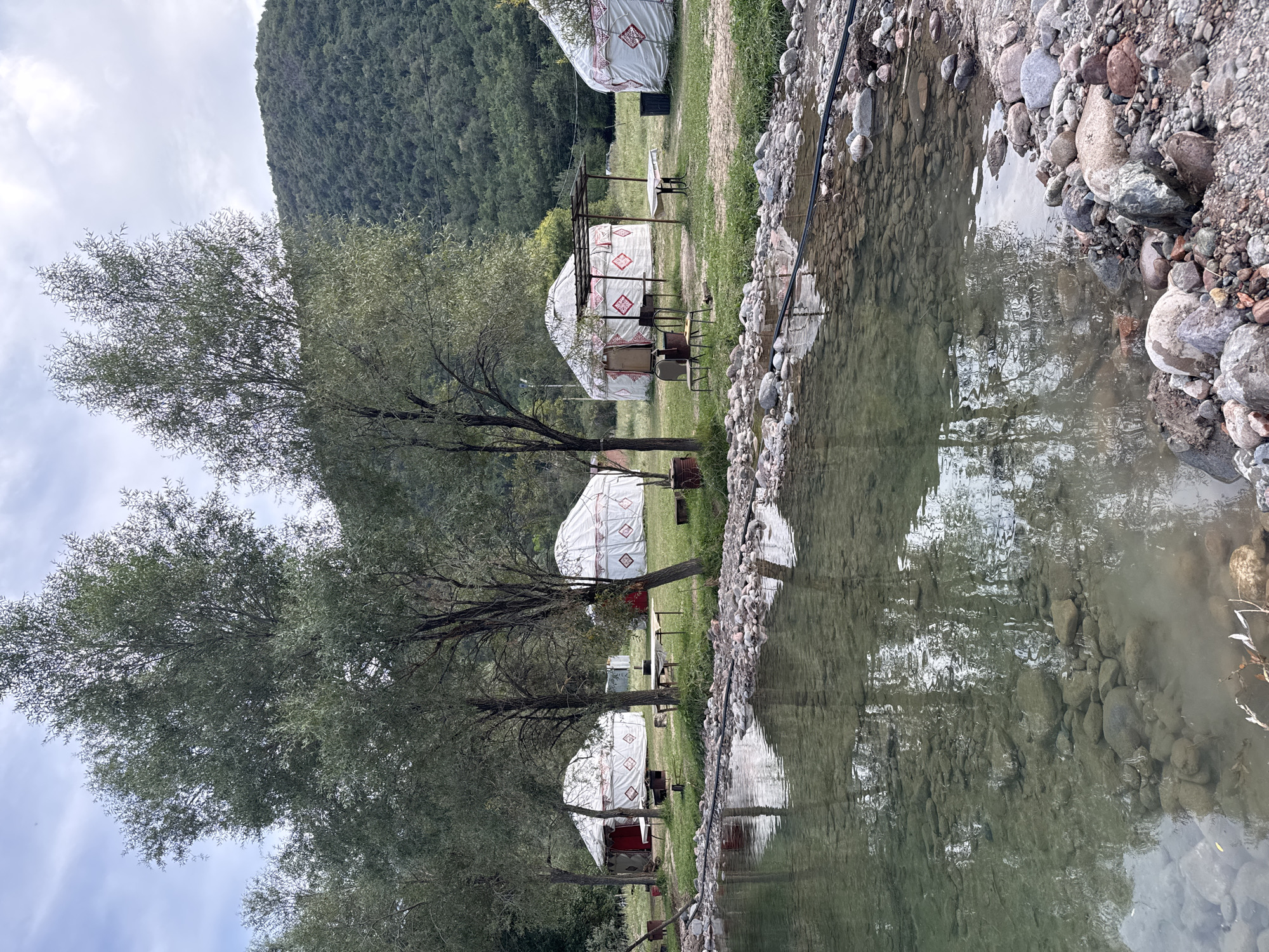 In Leskhoz, Kazakhstan, several traditional yurts are set up on a grassy area beside a clear, shallow body of water. The yurts are white with red geometric patterns and traditional embellishments. They are arranged in a semi-circle, with some tables and chairs placed between them, suggesting a communal or recreational area. Trees with green foliage are scattered around the yurts, providing shade and natural beauty. In the background, a lush, green, tree-covered hill rises, indicating a natural, possibly mountainous, landscape. The water in the foreground is calm and transparent, revealing the rocky bottom. The sky is overcast with scattered clouds. The overall scene depicts a peaceful and rustic retreat, characteristic of rural Kazakhstan.