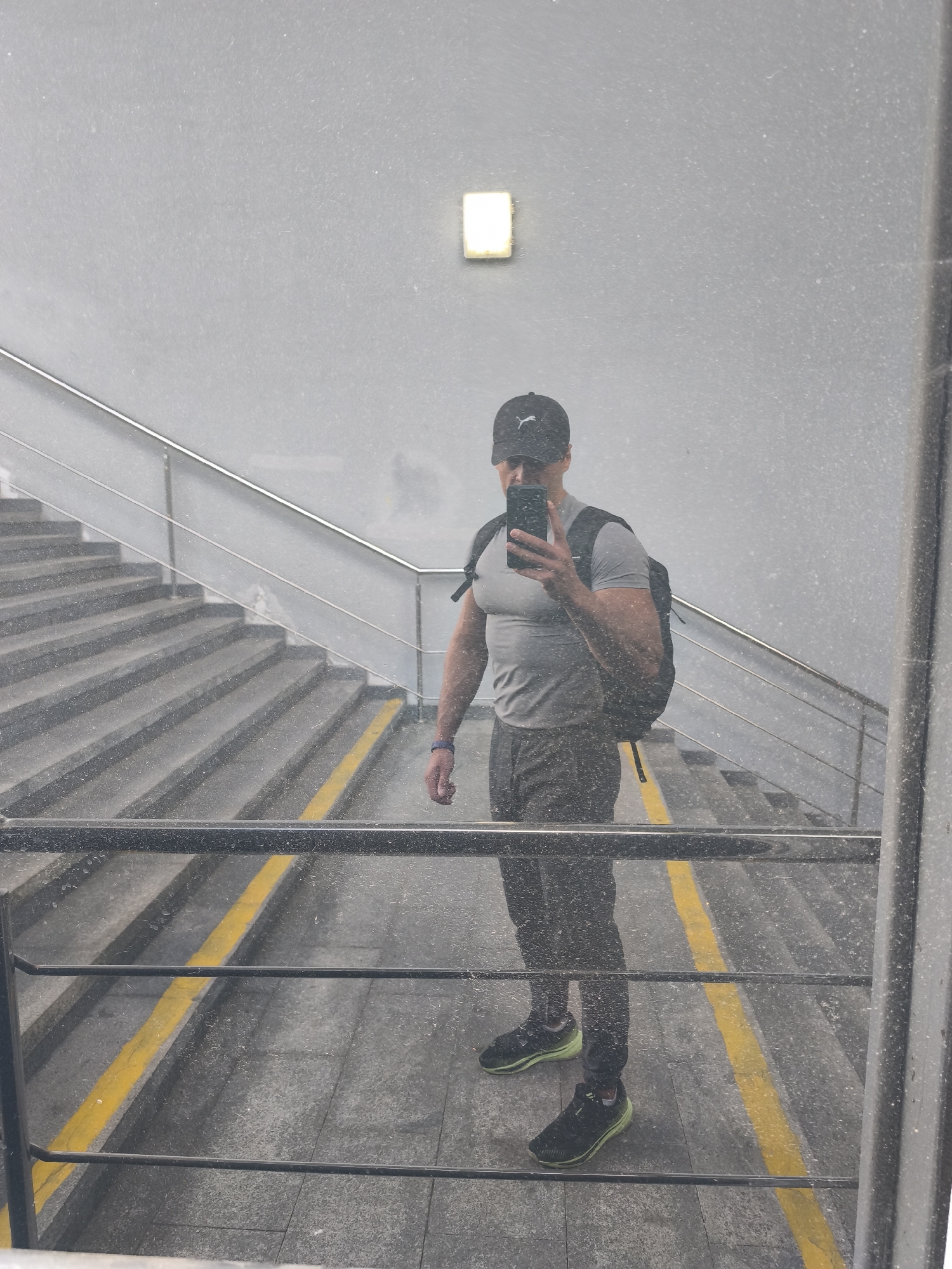 A man, wearing a grey t-shirt, dark pants, a black baseball cap, and black sneakers with light green accents, stands on a staircase. He is reflected in a mirror, holding a smartphone in his right hand to take a selfie. The man is also carrying a black backpack. The staircase is made of concrete with a metal railing. Yellow lines mark the edge of the steps. The background appears to be an indoor or covered area, with a grey wall and a light fixture. The overall impression is of a functional, perhaps public, space. The image is somewhat obscured by a layer of dust or smudges on the mirror's surface, creating a speckled effect throughout the photo. This suggests the mirror may be in a less-than-pristine condition, possibly in a transit area or a building undergoing maintenance. The lighting is neutral, and there are no strong indications of time of day or specific weather conditions, although the dust might hint at an indoor or sheltered environment. The man's expression is not clearly visible due to the angle and the reflection. No text is visible in the image.