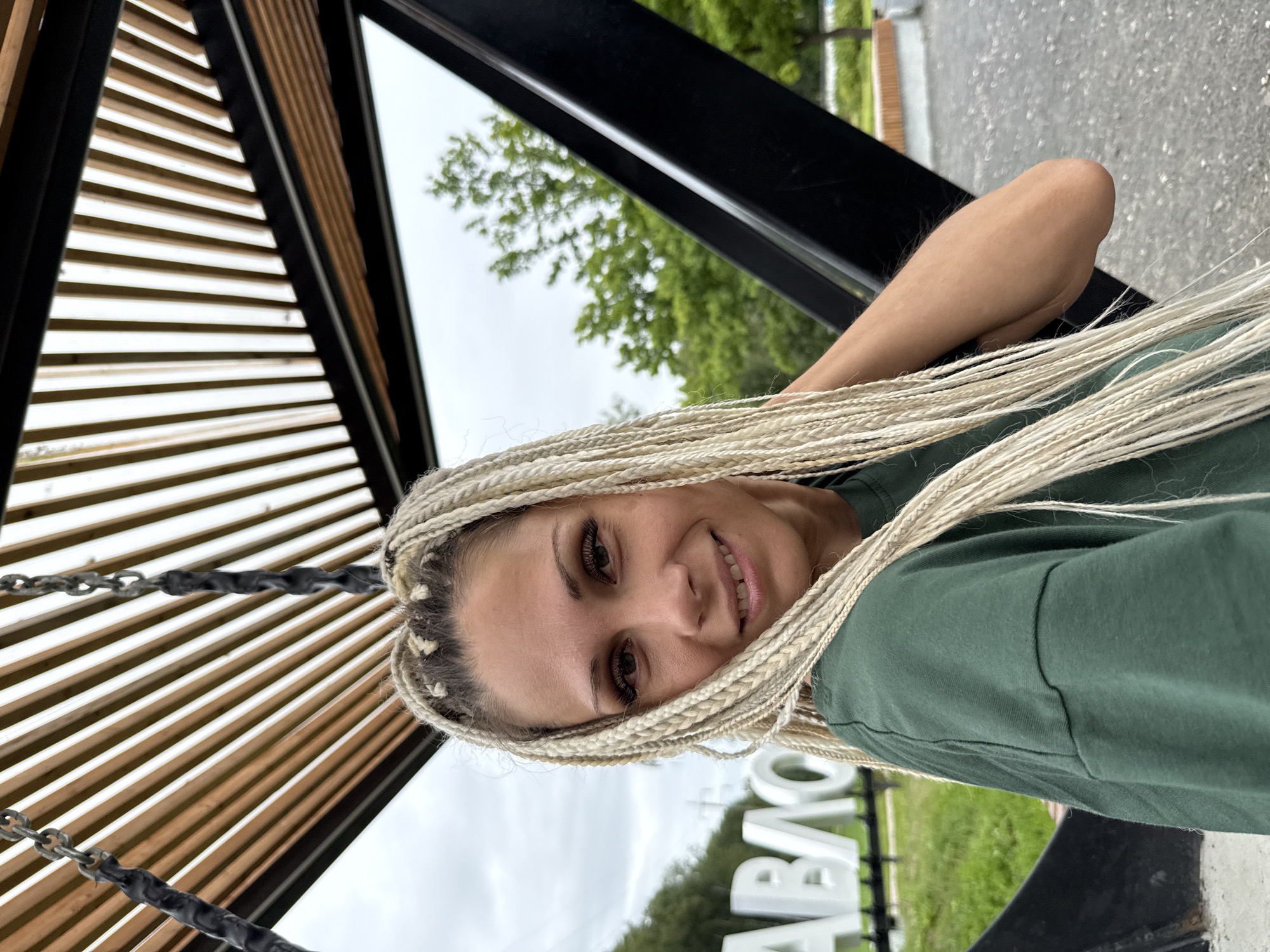 A young woman with blonde braided hair is pictured from the chest up, outdoors in what appears to be a public park or recreational area. She is wearing a dark green t-shirt and smiling at the camera. Behind her, large white letters spelling out "RUSANOVO" are visible, indicating the location. The background features trees and a cloudy sky, with a wooden slatted structure overhead.