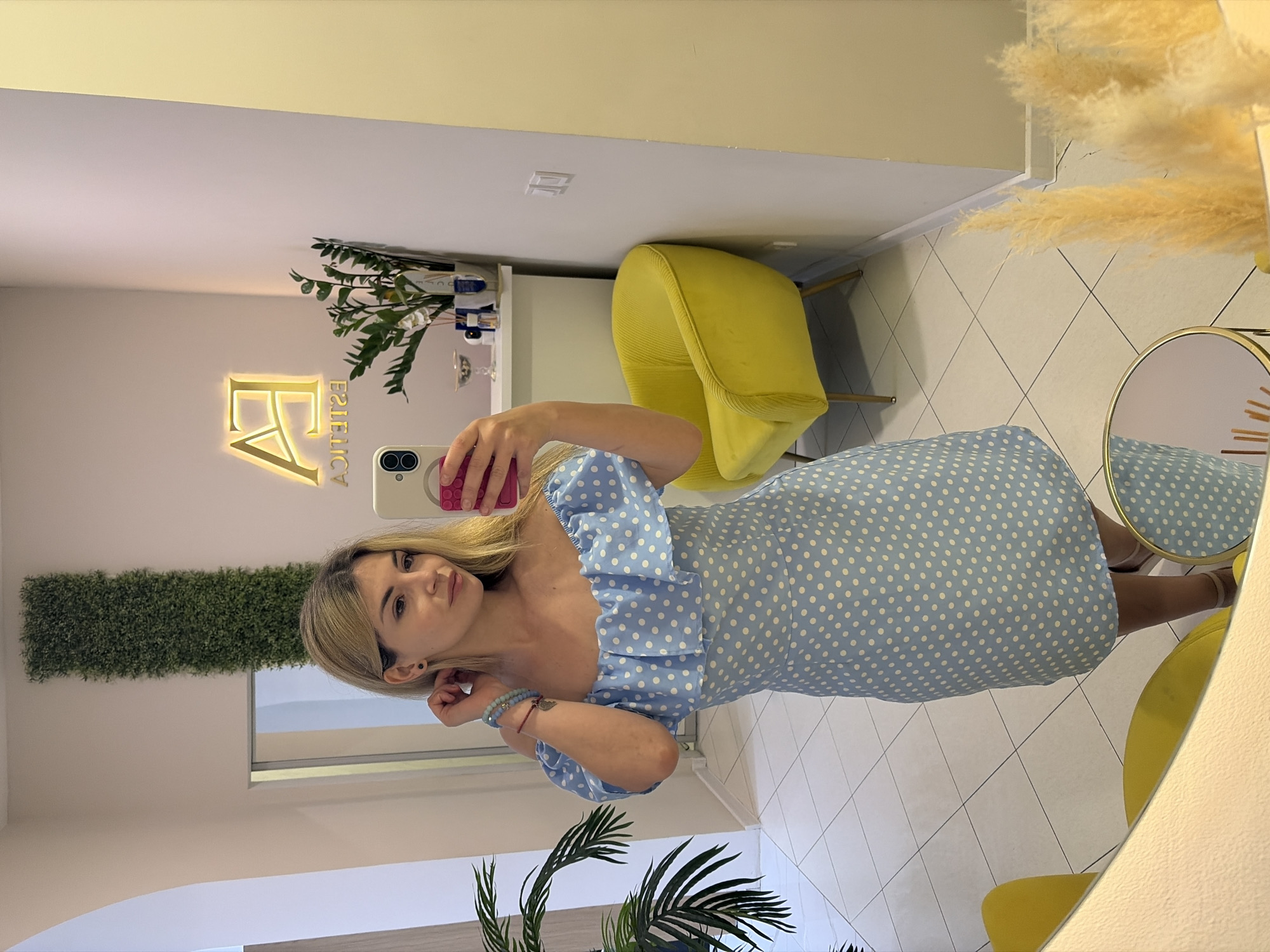 A young woman with blonde hair is captured in a mirror selfie within what appears to be a modern beauty salon or aesthetics clinic in Budva, Montenegro. She is wearing a light blue dress adorned with white polka dots and featuring ruffled details around the neckline and sleeves. Her left wrist displays multiple bracelets, and she holds a smartphone with a light-colored case and a pink grip, reflecting her image in the mirror.
The indoor setting is brightly lit, showcasing clean, light-colored walls and a tiled floor with a light square pattern and dark grout lines arranged diagonally. Prominently featured on the wall behind the woman is a luminous gold sign that reads "FA ESTETICA," indicating the nature of the establishment. To the left, a long, vertical strip of artificial green foliage decorates the wall.
Other elements in the room include a vibrant yellow upholstered chair, a table with various beauty products in bottles and jars, and a decorative plant with broad green leaves in the foreground. In the upper right corner, fluffy pampas grass adds to the decor, next to a partial view of a round mirror with a gold frame. The overall atmosphere is clean, contemporary, and inviting, suggesting a professional and well-maintained space.