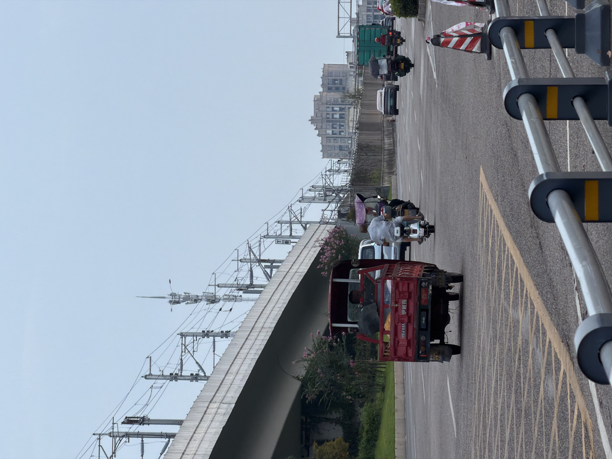 The image depicts a street scene in Jiangjin District, Chongqing, China, under an overcast sky. A multi-lane asphalt road dominates the foreground, marked with yellow and white lines. In the middle ground, various vehicles are in motion. A red, three-wheeled utility vehicle with a canopy is moving away from the camera, carrying at least one person in its cabin. To its right, several motorcycles and a scooter are also traveling. One scooter has a passenger with a purple canopy over their head. Further down the road, a green truck is visible.
The background reveals an elevated structure, likely an overpass for a railway or highway, supported by concrete pillars and adorned with numerous overhead wires and poles, indicative of electrical infrastructure. Beyond this, a large, multi-story building with a distinctive architectural style stands prominently. The area adjacent to the road on the left side features a grassy slope and some pink flowering bushes. On the right side, traffic cones with red and white stripes are placed along the roadside. The overall atmosphere suggests a moderately busy urban environment during daylight hours.