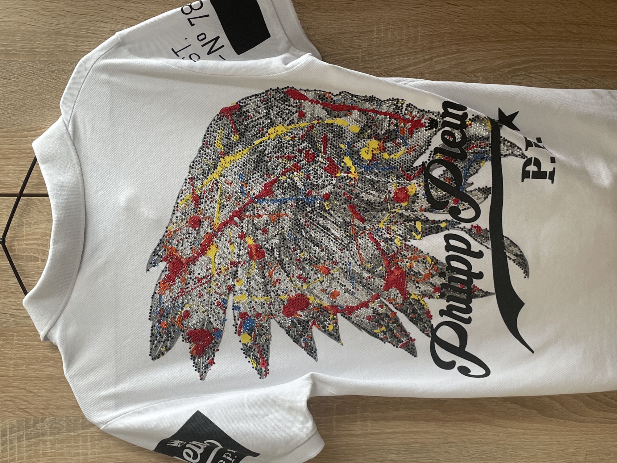 A white Philipp Plein T-shirt is lying on a wooden surface. The shirt has a large graphic design on the front. The design features a stylized wing made of small, possibly reflective, elements in shades of grey, red, yellow, and blue. The Philipp Plein logo is printed in a large, stylized black font next to the wing. The letters "P.P." are also printed on the shirt. There is a black square label on the upper shoulder area of the shirt and another black square label on the lower left corner of the shirt. There is a black hanger partially visible near the top left corner. The scene is indoors. The location in the city of Sofia, Bulgaria cannot be determined from the image.