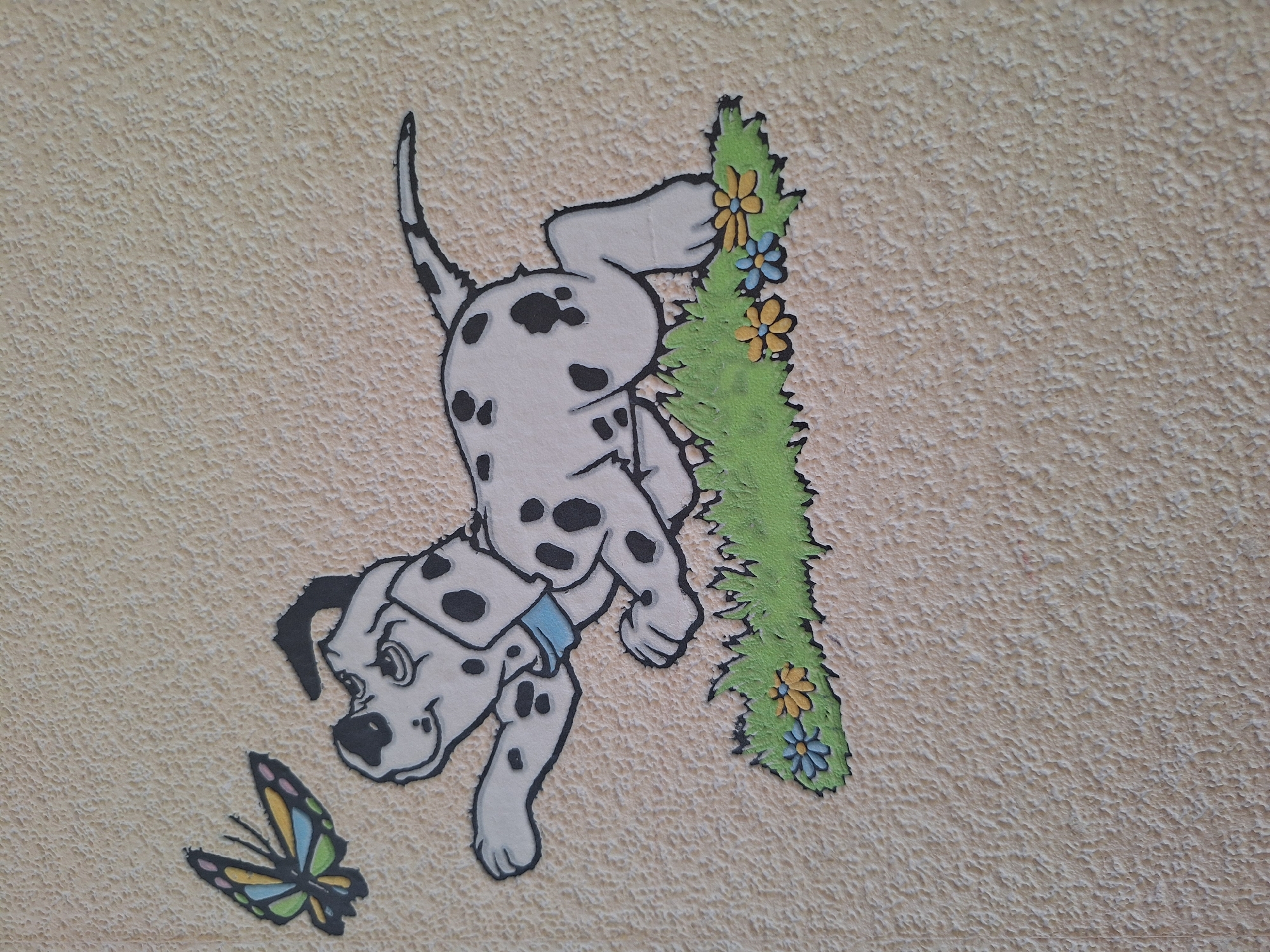This is an image of a wall mural featuring a Dalmatian puppy and a butterfly. The puppy is white with black spots and is depicted in a playful pose, with its front paws extended and its body low to the ground. It is wearing a blue collar. Above and to the left of the puppy is a colorful butterfly with its wings spread. The lower part of the mural shows a patch of green grass with small yellow and blue flowers. The background of the mural is a textured, light beige surface, likely a wall. The mural appears to be a decorative element, possibly in a child's room or a public space. There is no identifiable information in the image to pinpoint its exact location within the city of Varna, Bulgaria.