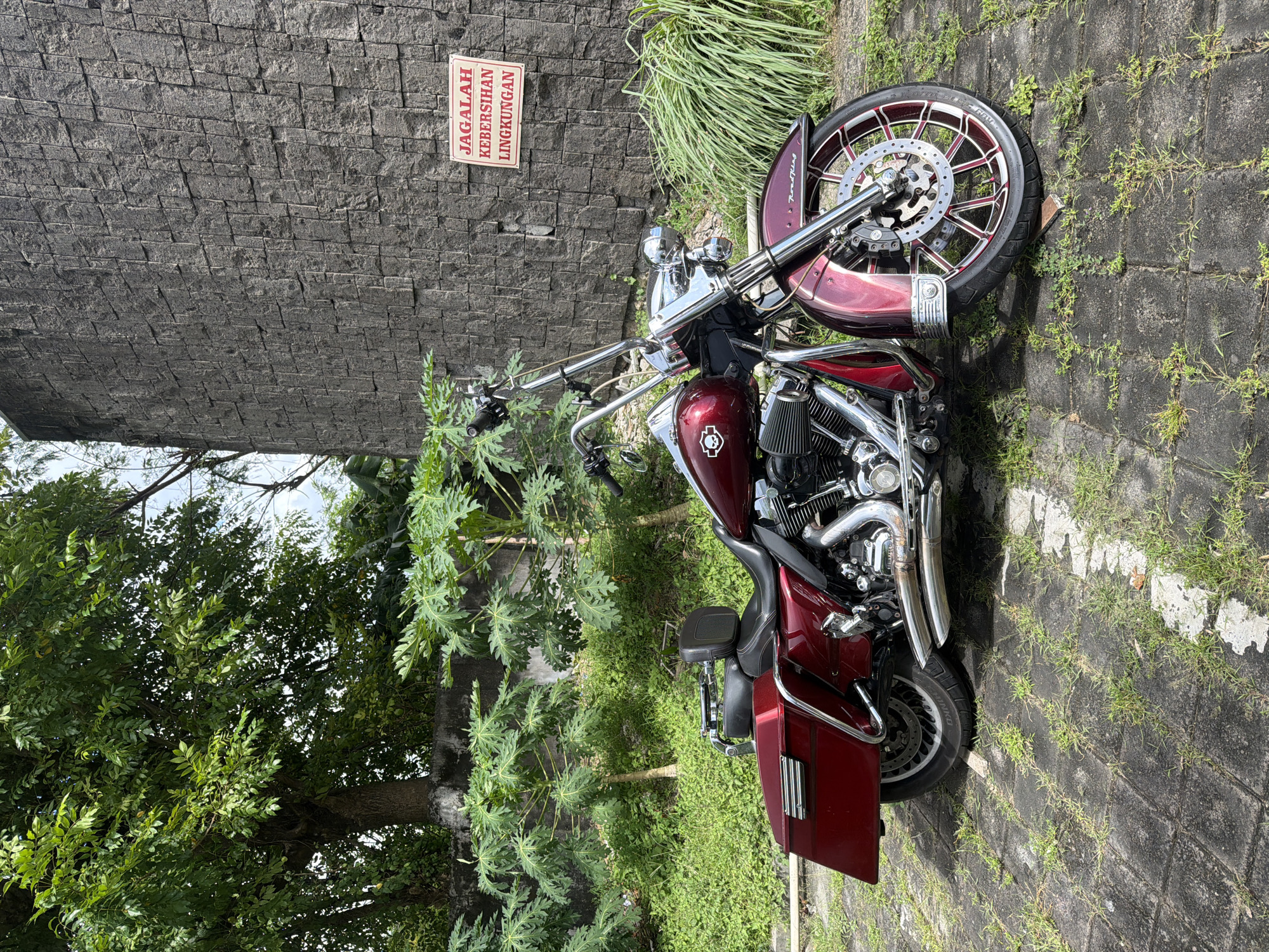 The image shows a red Harley-Davidson motorcycle parked near a stone wall and some greenery. A sign on the wall is in Indonesian. The surrounding environment consists of trees, grass, and brick pavement. The location is Unknown, Unknown.