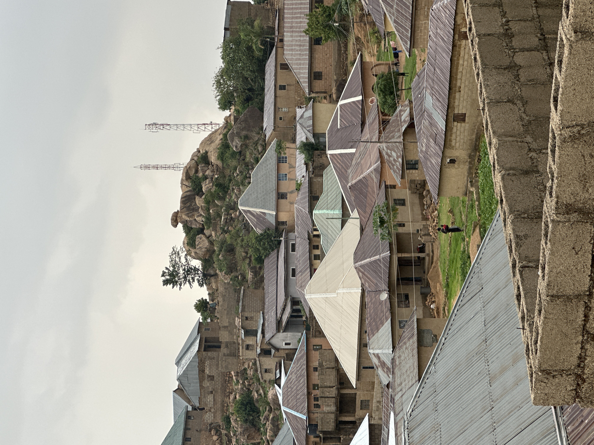 The image displays a sprawling, densely built settlement nestled among rugged, rocky hills under an overcast sky. Numerous houses, primarily single-story, are constructed from light brown concrete blocks. Their roofs are predominantly made of corrugated metal sheets, showing varying shades of grey, brown, and some with hints of faded green, indicating age and weather exposure. Many roofs exhibit rust and wear.
The topography is defined by prominent rocky outcrops that rise in the background and are integrated into the urban fabric, with some buildings constructed directly on their slopes. Sparse green vegetation, including small trees and patches of grass, is visible among the buildings and rocks. Two tall, slender metal communication towers stand atop one of the highest rocky formations, silhouetted against the uniformly grey sky.
Evidence of daily life is present, with a few human figures discernible. One person, dressed in dark clothing with a red backpack, is seen standing or walking in a grassy area between houses. Another smaller, dark figure is visible near laundry hanging outside a building further back. In the immediate foreground on the right, a rough concrete block wall suggests an ongoing construction or a boundary overlooking the settlement.
The sky is entirely overcast, casting diffused and even light across the scene, with no discernible shadows. This indicates a cloudy day, potentially early morning or late afternoon. The architectural style, materials, and the integration of housing with the rocky landscape are consistent with the provided context of Jos, Nigeria. There is no visible text in the image.