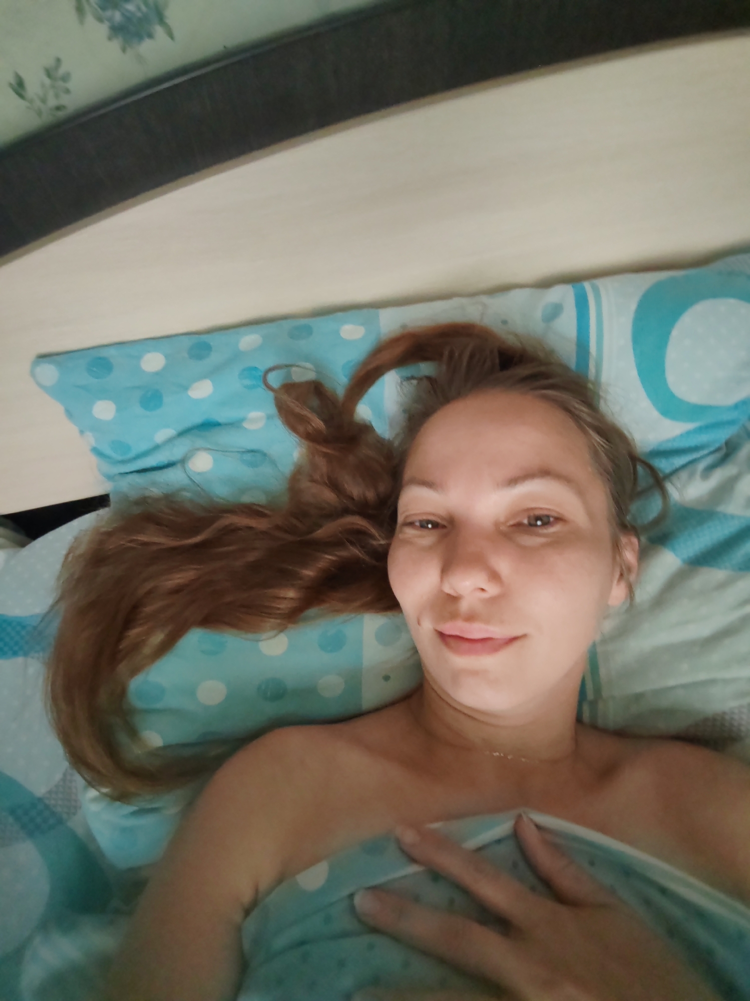 The image shows a young woman lying on a bed in what appears to be a bedroom. She is looking at the camera and has long, reddish-brown hair. She is partially covered by a light blue blanket with white polka dots. The bedding also features a light blue and white pattern. The background shows a headboard and what seems to be a wall with a decorative pattern. The lighting suggests it is daytime. This scene is taking place in a residential setting within the city of Chukhlem, Russia.