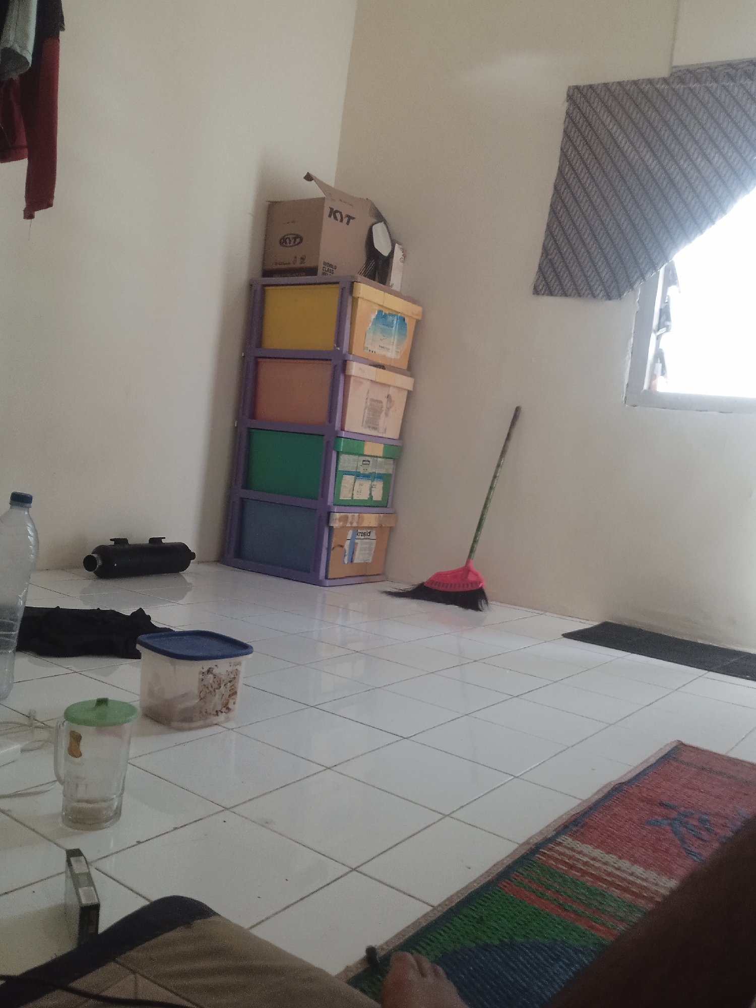 The image depicts an interior scene, likely a room, with white tiled flooring. In the foreground, a person's leg and foot are visible resting on a woven mat. To the left, a clear glass with a green lid sits next to a plastic container with a blue lid. Further left, a large clear plastic bottle is partially visible. A black cylindrical object is on the floor against the wall.
Dominating the mid-ground is a tall, multi-colored plastic storage unit filled with boxes. The unit has shelves colored purple, yellow, orange, green, and blue. On top of the unit are cardboard boxes, one with "KYT" visible. A broom with a pink head leans against the wall near the storage unit.
In the background, a window with a patterned blind is visible. The overall impression is a somewhat cluttered but functional living space. The presence of a woven mat and the general aesthetic suggest this could be a typical home interior in Slorok, Indonesia.