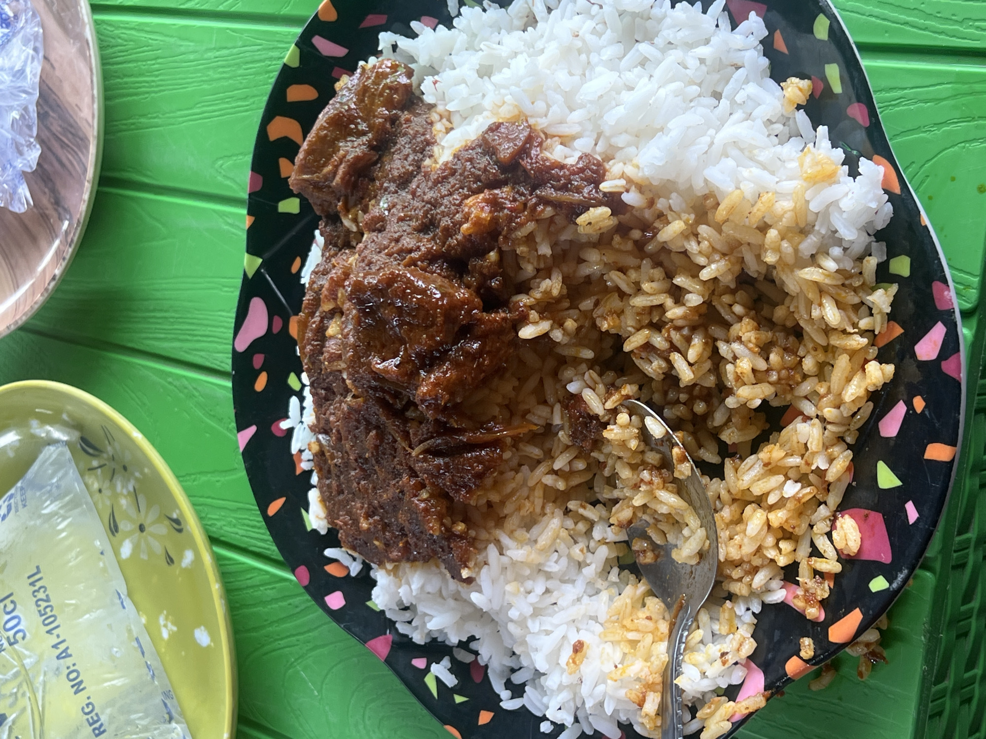 A high-angle shot depicts a plate of food on a green, textured surface. The plate is black with a scattered confetti pattern of pink, orange, green, and white. The food consists of a portion of white rice and a portion of brown stew with meat. A spoon rests on the rice. In the lower left corner, a yellow plate contains a sealed plastic bag of water. The image was taken in Jalingo, Nigeria.