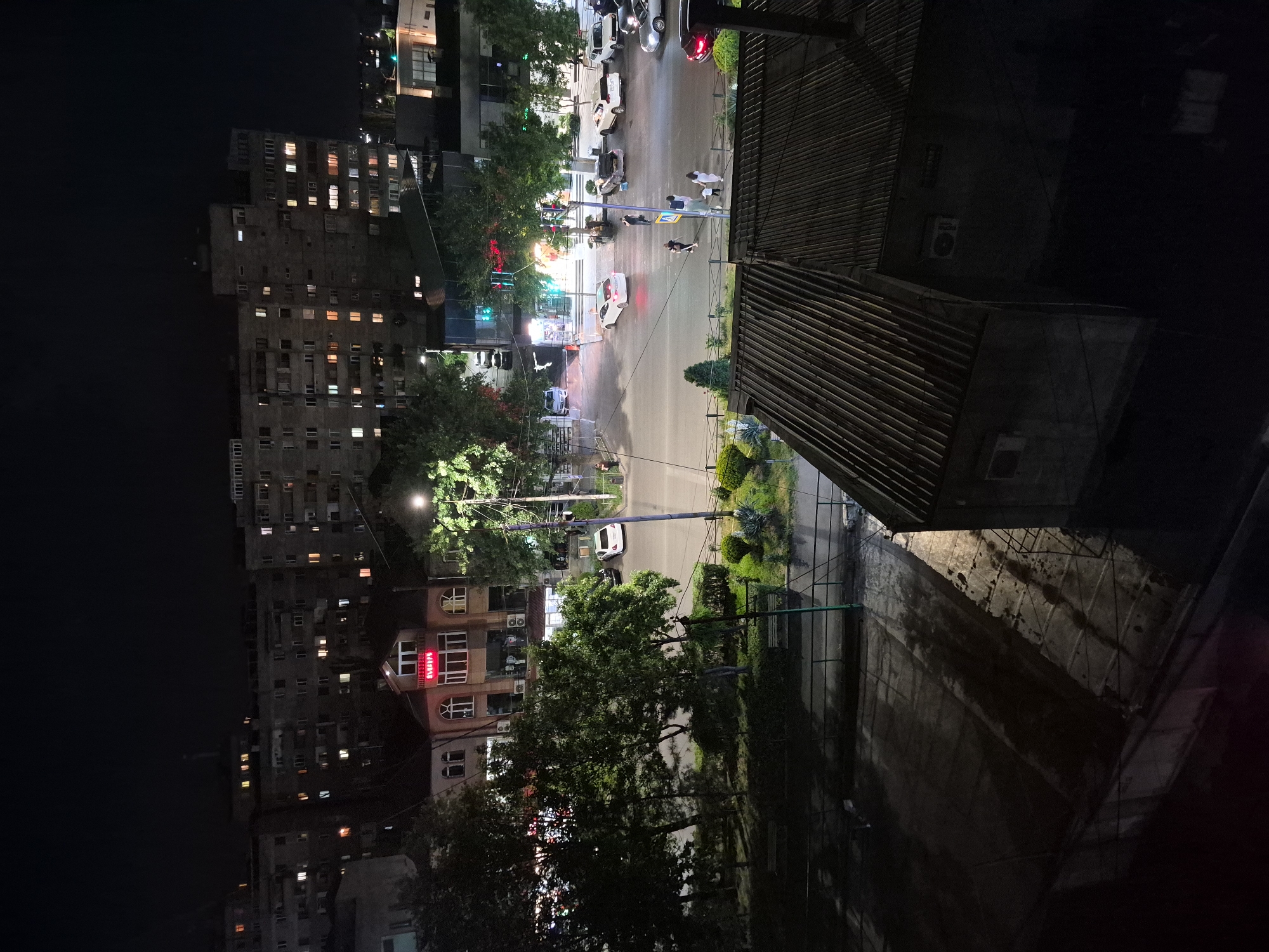 This night-time image, captured from an elevated viewpoint in Yerevan, Armenia, depicts a bustling urban street scene. The dark sky indicates the time of day, with various artificial lights illuminating the setting.
On the left side, a tall, multi-story residential building dominates, its facade featuring numerous windows, many of which are lit, suggesting ongoing activity within. Below and to the right of this large building, another commercial structure is visible, distinguished by a prominent, brightly lit red sign with white Armenian text that reads "ԲԱՆԿԻ" (BANKI), identifying it as a bank.
The central part of the image features a street and an intersection. Several vehicles, including both parked and moving cars, are present; a white car is notably visible near a traffic light. The street surface appears somewhat reflective, possibly due to dampness or light reflections. Traffic lights, displaying both red and green signals, regulate movement at the intersection. Multiple pedestrians are seen on the sidewalks and crossing the street, contributing to the city's evening activity. The street is flanked by trees with green foliage and illuminated by streetlights.
In the immediate foreground on the right, a distinctive slatted roof structure is prominent, along with a dark, textured wall below it. An air conditioning unit can be spotted on an adjacent building surface in the upper right section of the image. The scene conveys a typical, active city atmosphere after dusk.