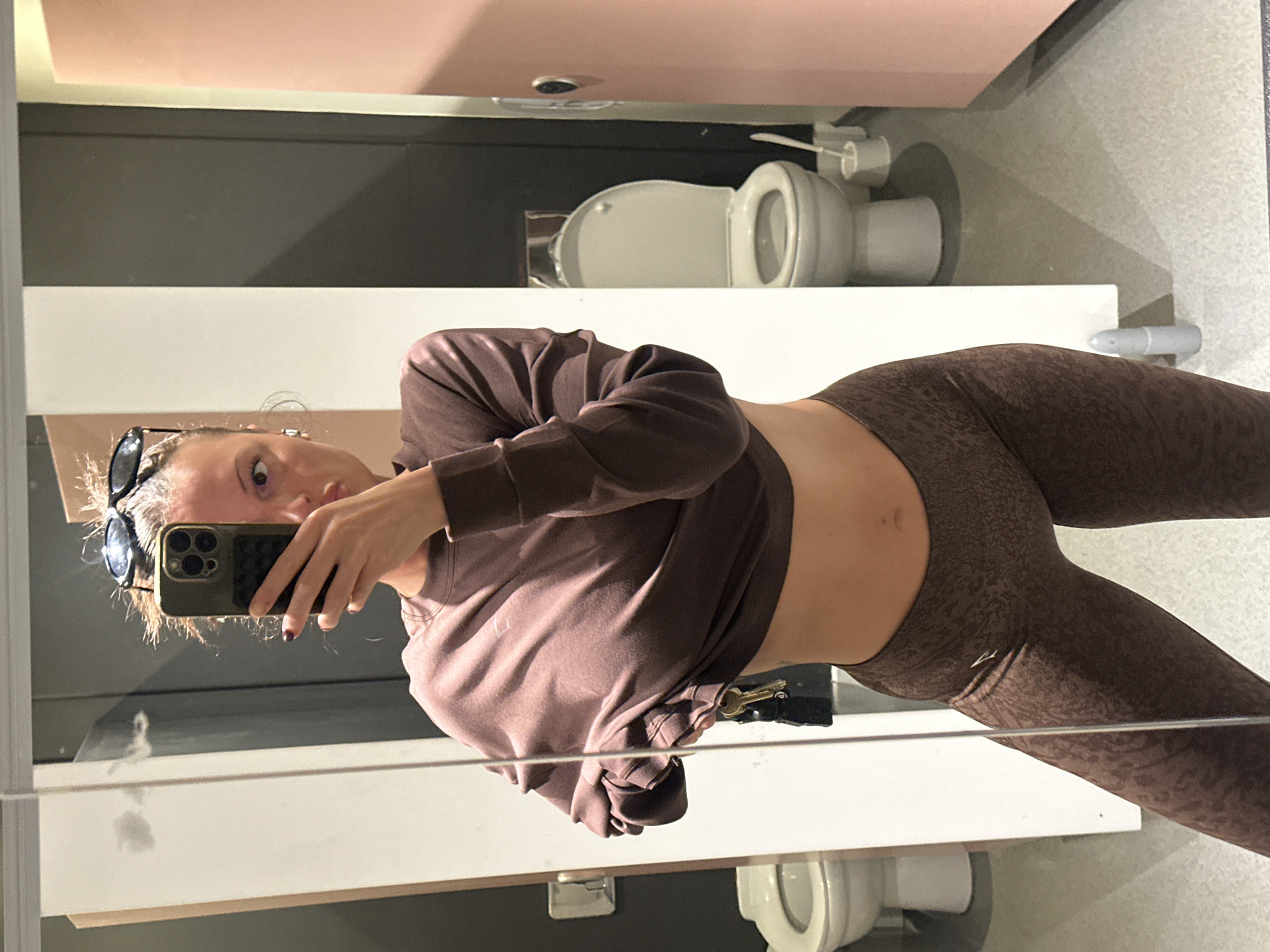 A person, likely female, is taking a mirror selfie in what appears to be a restroom in Tonbridge and Malling, United Kingdom. The image is taken from a slightly elevated angle, capturing her reflection from the chest down, and is rotated 90 degrees clockwise from standard orientation.
She is holding a dark-colored smartphone, identifiable as an iPhone by its distinct camera module, to take the photo, which partially obscures her face. Her light brown hair is pulled back, and a pair of dark sunglasses rests on top of her head. Her expression is neutral, with her eyes wide open.
The person is dressed in a cohesive outfit consisting of a dark brown, long-sleeved cropped top that reveals her midriff, paired with matching dark brown patterned leggings. A single gold-colored key attached to a keyring is visible, hanging from her waistline.
The background, seen through the mirror's reflection, depicts a clean and modern bathroom environment. A white toilet with its lid raised is clearly visible in the lower portion of the reflection. Above the toilet, a toilet paper holder is mounted on the wall. The walls behind the person are segmented horizontally with distinct color blocks: a light pink or peach hue at the top, a dark grey band in the middle, and a white section below that. The floor appears to be a light-colored tiled or linoleum surface. The lighting is artificial, indicating an indoor setting. No discernible text is visible within the image.
