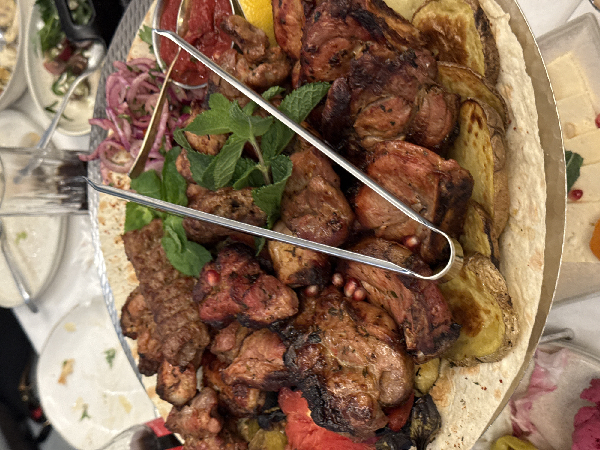 An overhead shot captures a large, circular platter filled with a generous spread of food, typical of an Armenian khorovats (barbecue) or shish kebab. The centerpiece is a substantial pile of grilled meat, likely lamb or pork, characterized by its well-browned and slightly charred exterior. Interspersed with the meat are golden-brown grilled potato wedges.
The dish is vibrantly garnished with fresh green mint sprigs, thinly sliced and possibly sumac-dusted red onions, and scattered ruby-red pomegranate seeds, adding a burst of color and flavor. A bright yellow lemon wedge is also visible, ready to be squeezed over the meal. The entire ensemble is served atop a bed of thin, soft flatbread, resembling lavash, which lines the platter.
A pair of silver metal serving tongs rest on the food, indicating it is prepared for communal serving. In the blurred background, parts of other dishes, bowls, and cutlery on white surfaces suggest a larger meal or banquet taking place indoors, possibly in a restaurant or during a family gathering in Yerevan, Armenia. The lighting illuminates the rich textures and appealing colors of the food.
