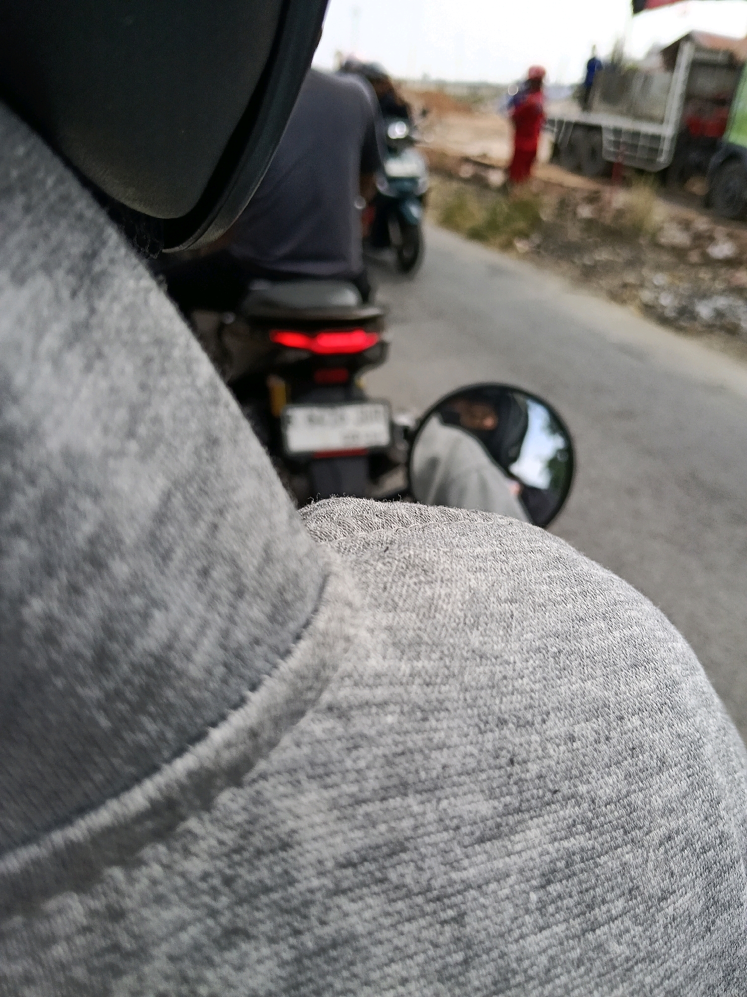 A close-up, first-person perspective shot shows the shoulder and back of someone wearing a grey hoodie, with the hood up, obscuring their head. The person is on a motorcycle, and the shot focuses on their clothing. In the midground, another motorcycle is visible with a rider wearing a dark shirt, and its red taillight is illuminated. Further in the background, on the right side of the road, a truck is parked. There are several people near the truck; one person is wearing a red outfit, and another appears to be wearing blue. The road appears to be asphalt, with a dusty shoulder on the right side, indicating a potentially rural or construction area. The lighting suggests it is daytime, with overcast skies. The scene is consistent with a street in Pajang, Indonesia, likely a commercial or industrial area due to the presence of a truck and road construction materials.
