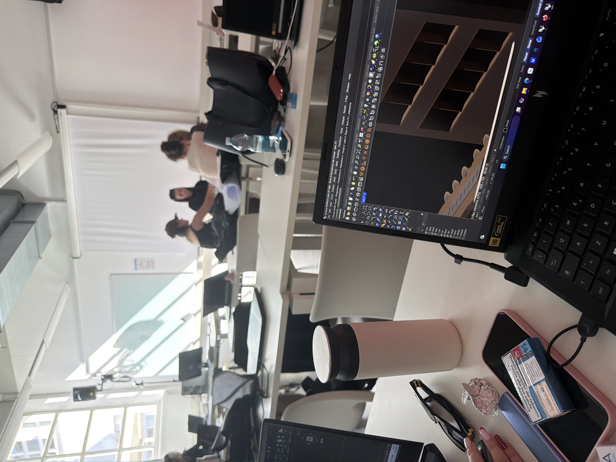 The image shows a desk in a bright, modern interior, likely in a co-working space or a university design lab in Milan, Italy. A laptop dominates the foreground, displaying a 3D rendering software interface with a model of shelves. The desktop area also includes a water bottle, glasses, a phone, a small piece of crumpled foil, and a package of tobacco products.
In the background, three people are seated at desks, engaged in what appears to be a design session or class. The overall impression is of a creative, collaborative workspace.
