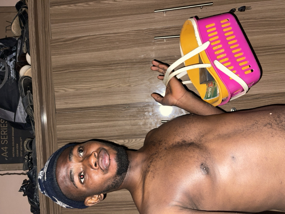 The image depicts an adult male standing indoors, identified as being in Muchiya, Nigeria. The male is bare-chested, exhibiting dark skin and visible dark body hair on his chest and abdomen. He wears a dark blue knit beanie with a lighter pattern. He has a short beard and full lips, slightly parted. His gaze is directed upwards and to the viewer's right; his left eye shows a distinct red reflection, consistent with a camera flash effect.
In his left hand, he holds a vibrant multi-colored plastic basket. The basket's main body is hot pink with vertical slatted sides, featuring a yellow/orange rim and interior, and two white plastic handles. Visible inside the basket are what appear to be several green and white currency notes and a dark, rectangular object.
The background consists of a wooden wardrobe or cabinet with vertical wood grain patterns. On the right side of the wardrobe, two vertical metallic handle bars are present, and a set of keys hangs from a small fixture. Resting on top of the wardrobe are various items including multiple pairs of sandals or slippers, dark fabric or clothing, and a cardboard box partially labeled "A4 SERIES" and "NATURAL COLOUR ENHANCEMENT," with additional text "DTS VIRTUAL:X" also visible. The scene is illuminated by artificial light.