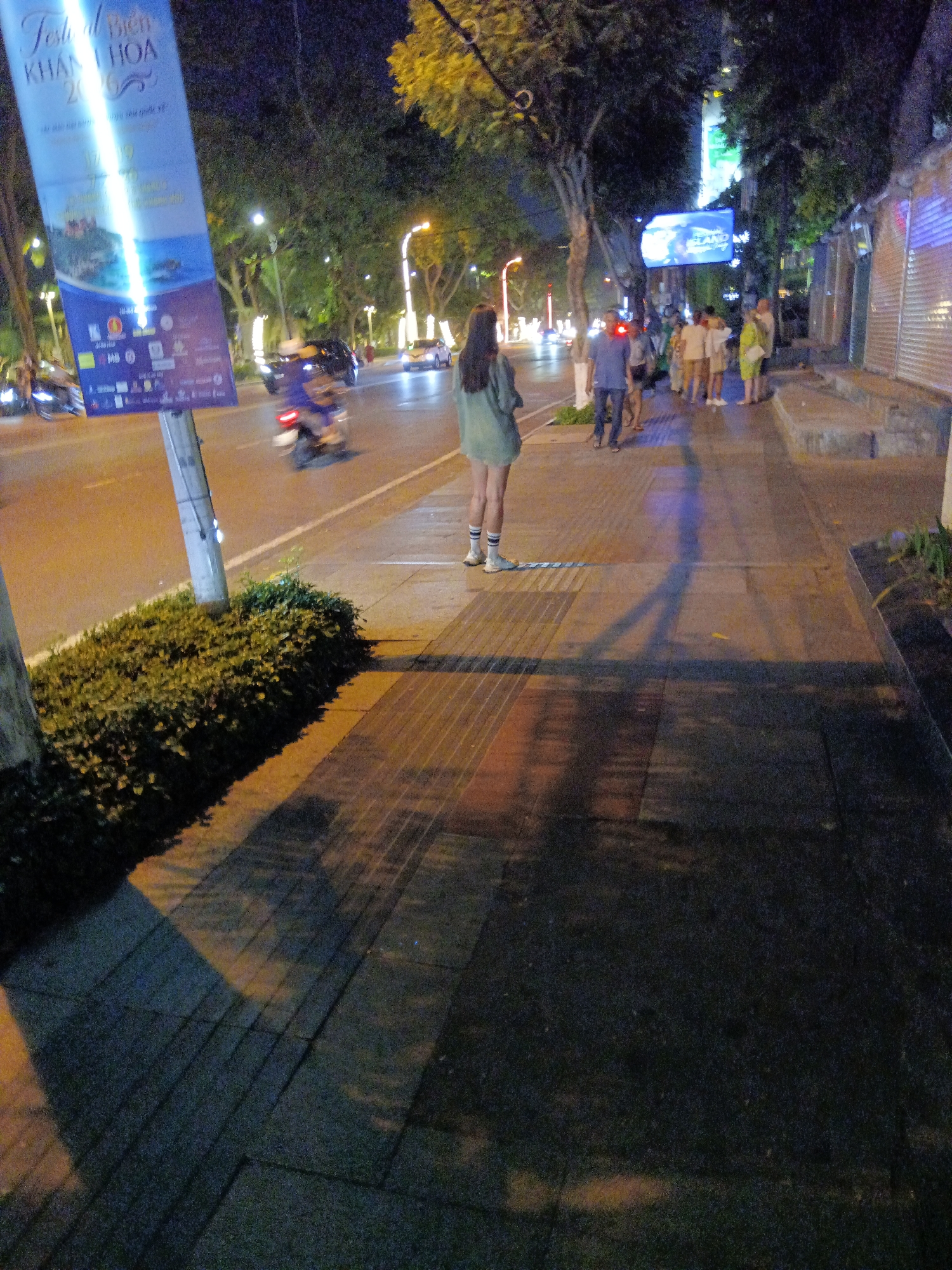 The image captures a street scene at night in Nha Trang, Vietnam, likely near a promenade or bustling area. A large, illuminated banner on the left advertises "Festival Bien Khanh Hoa 2023." In the foreground, a young woman stands on a sidewalk, facing away from the camera, wearing a light green oversized shirt and dark shorts, with white socks and shoes. She is positioned in front of a neatly trimmed hedge bordering the sidewalk.
The sidewalk is paved with rectangular tiles in varying shades of orange and grey, with textured strips indicating a pedestrian crossing or tactile paving. To the right of the sidewalk, a low concrete ledge and a dark-colored wall suggest a building facade, possibly shops or businesses, with corrugated metal shutters visible.
Across the street, a busy road is visible with traffic, including a moving motorbike and several cars. Streetlights illuminate the scene, casting long shadows on the sidewalk. In the background, a group of people are gathered on the sidewalk, some embracing, and trees line the street. The overall ambiance is that of a lively evening in a coastal city.