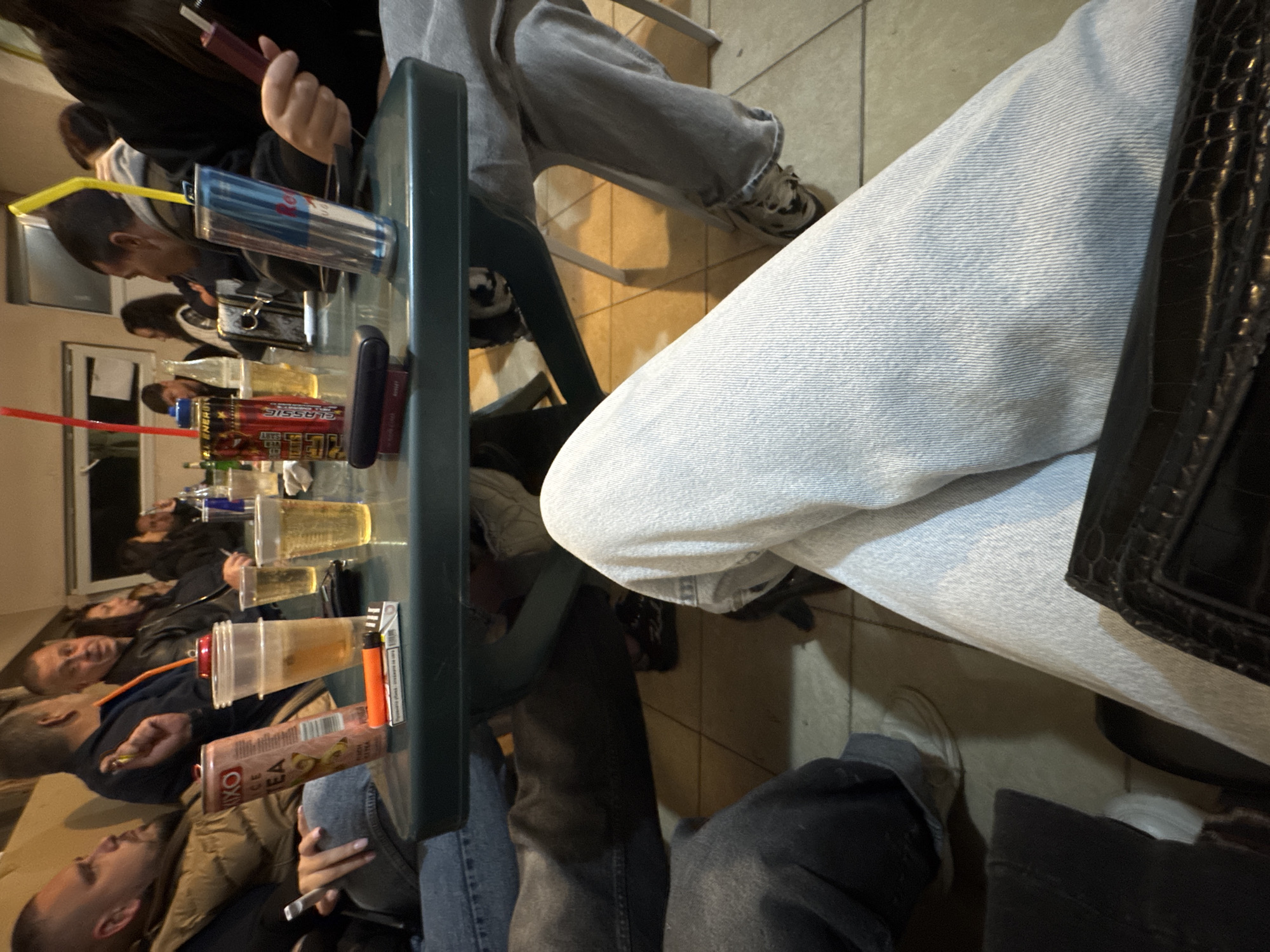 The image depicts a group of people seated at a table, likely outdoors, based on the ambient lighting. Several individuals are visible, engaged in conversation and holding various drinks and items. There's a table covered with various beverage containers, including cups, cans, and bottles. The attire suggests casual clothing. The location, Topolovets, Bulgaria, is stated, but without further visual references in the image, no specific landmarks or identifying features are visible to confirm the location within the city. The lighting is somewhat dim, indicating it could be evening or a dimly lit indoor setting. The environment shows basic tiled flooring. Interactions seem social, based on the proximity and assumed engagement among the individuals.