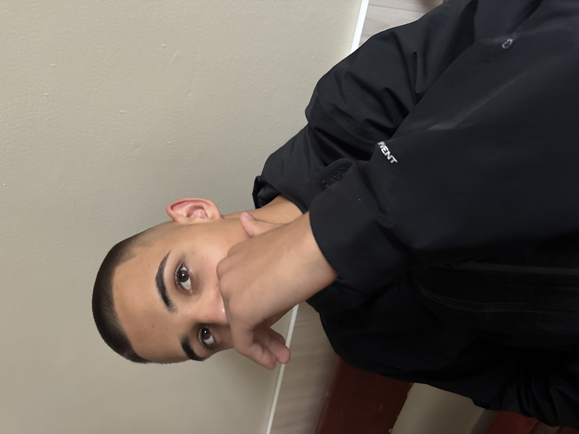 An upward-oriented (portrait mode, rotated) medium close-up shot features a young individual, likely male, looking directly into the camera. The person has short, dark hair styled in a clean fade or buzz cut, prominent dark eyebrows, and expressive brown eyes. Their skin tone is olive.
They are wearing a black jacket or coat, with part of one sleeve visible, revealing white text that reads "MENT." One hand is raised, with fingers partially covering the lower part of their face, from the nose down, obscuring their mouth. The pose suggests a moment of contemplation, shyness, or a casual gesture.
The background consists of a plain, light beige or off-white wall, which occupies most of the space behind the person. In the bottom right corner of the frame (when correctly oriented, it would be the bottom right of the image), a small section of a dark red or brown surface, possibly a floor or furniture, is visible. The lighting is soft and even, suggesting an indoor setting.