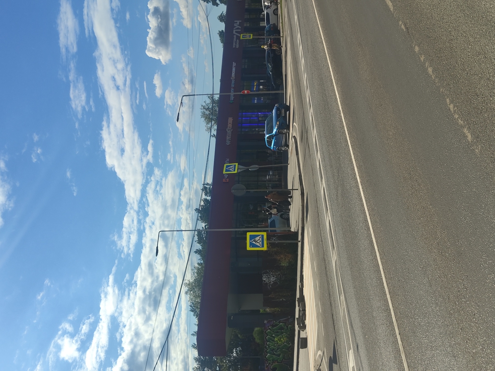 A sunny day view shows a commercial building complex in Ramenskoye, Russia, featuring a red overhang and a facade with several storefronts. Various signs are visible, including "ФРУКТЫ ОВОЩИ" (Fruits Vegetables), "ЗДОРОВЬЕ АПТЕКА СЕРВИСМАН" (Health Pharmacy Serviceman), and "ТОРГОВЫЙ ЦЕНТР ЛУЧ" (Shopping Center Luch). Pedestrian crossing signs with yellow backgrounds and a pictogram of a person walking are present on the sidewalk. A blue SUV is parked in front of the building, and several other vehicles are visible on the street. The foreground displays a paved road with lane markings and a crosswalk. Trees line the background behind the building.
