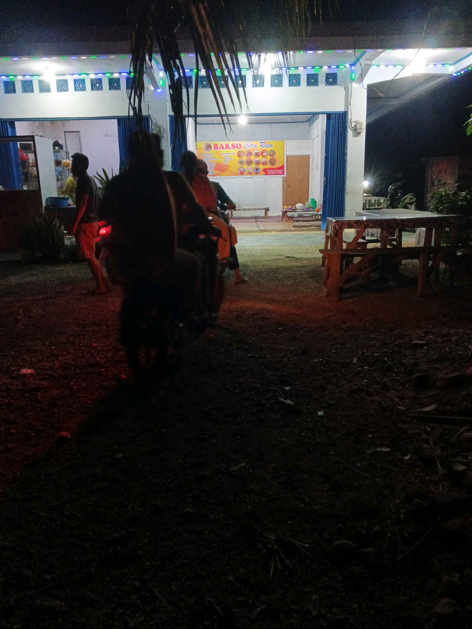 In Brangan Mulya, Indonesia, at night, a food stall identified by a sign reading "BAKSO Citra Rasa" is visible. The stall is illuminated by string lights and a fluorescent light above the entrance. Several people are present. In the foreground, a person on a motorbike is partially obscured by darkness. To the left, two individuals are standing near a counter. A wooden table and benches are set up outside the stall for customers. The ground is covered in gravel and dirt.