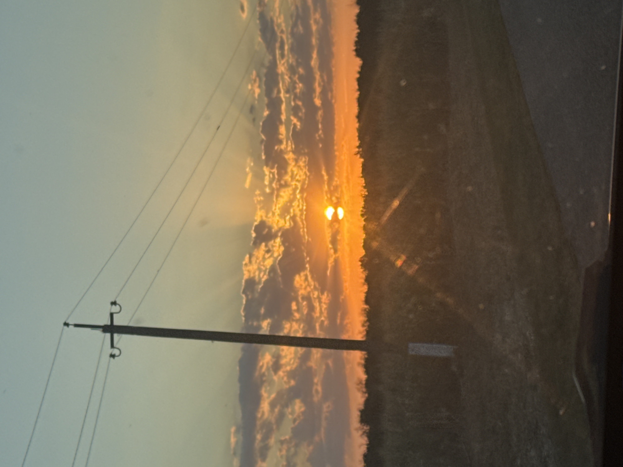 The image depicts a sunset over a rural landscape near Vostochny, Russia. A tall utility pole stands prominently on the left, with power lines extending horizontally across the frame. The sun, a bright orange orb, is setting behind a line of dark trees and vegetation in the distance, casting dramatic rays of light through scattered clouds. The sky transitions from a pale blue at the top to warm oranges and yellows near the horizon. In the foreground, a dark, sloped surface, possibly a road, is visible. The overall impression is one of a serene, natural setting illuminated by the warm glow of a setting sun.