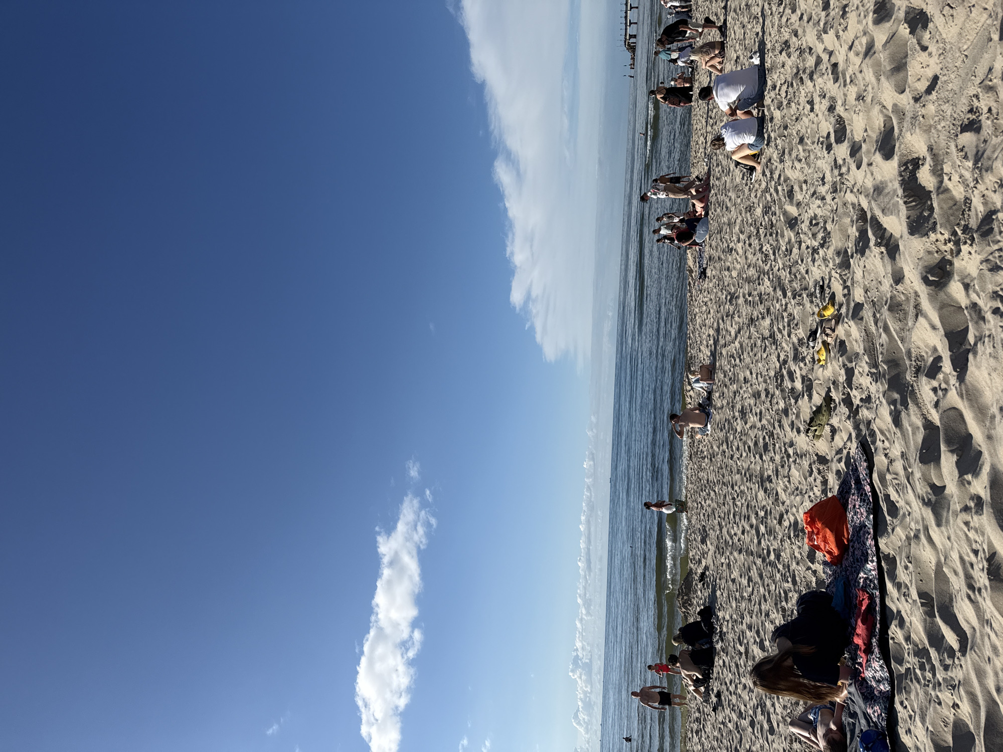 The image depicts a sunny day at the beach in Międzyzdroje, Poland. The foreground is dominated by sand dunes, with several groups of people relaxing on blankets and towels. Some individuals are seated, while others are lying down. Further back, closer to the water, a larger crowd of people are gathered, with some wading in the shallow waves of the Baltic Sea. The sea is calm with small waves gently lapping the shore. In the distance, a pier extends out into the water, and several boats are visible on the horizon. The sky is a bright blue with scattered white clouds. The overall scene conveys a sense of leisure and summer vacation.