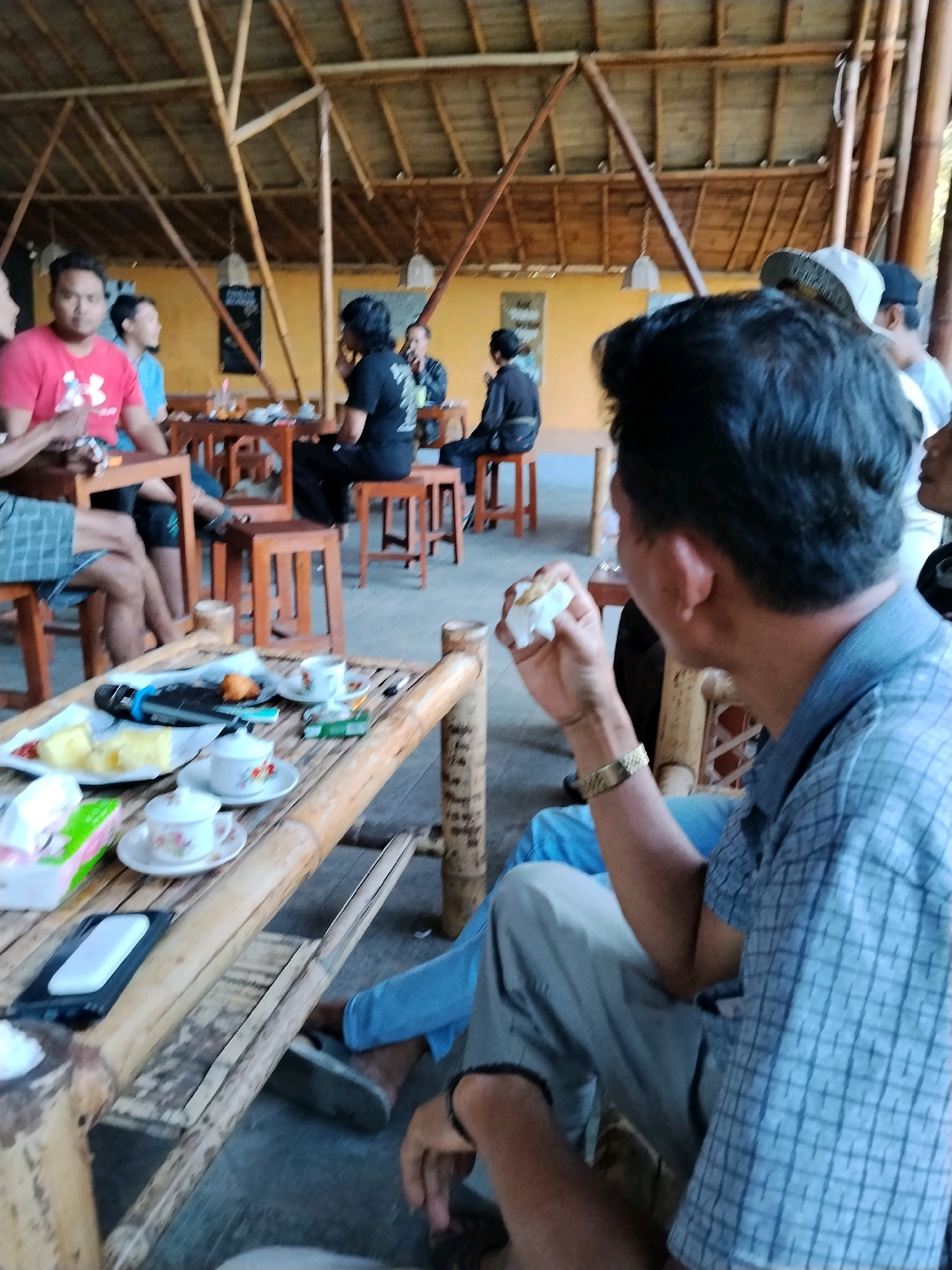 The image depicts a group of people gathered in what appears to be an outdoor seating area with a bamboo-themed structure. The setting suggests a casual dining or social gathering spot.
In the foreground, a man wearing a blue checkered shirt and light-colored pants is seated, holding a piece of food in his right hand. He is looking towards the right side of the frame. A bamboo table is positioned in front of him, laden with items including plates of food, cups and saucers, a pack of cigarettes, a microphone, and a mobile phone. A woman in a pink t-shirt is visible in the background, seated at a table. Other individuals are present in the background, some seated at tables and others standing, engaged in conversation or eating. The environment features a bamboo ceiling and yellow walls. The image does not provide specific indicators to pinpoint its location within the city of Pekalongan, Indonesia.