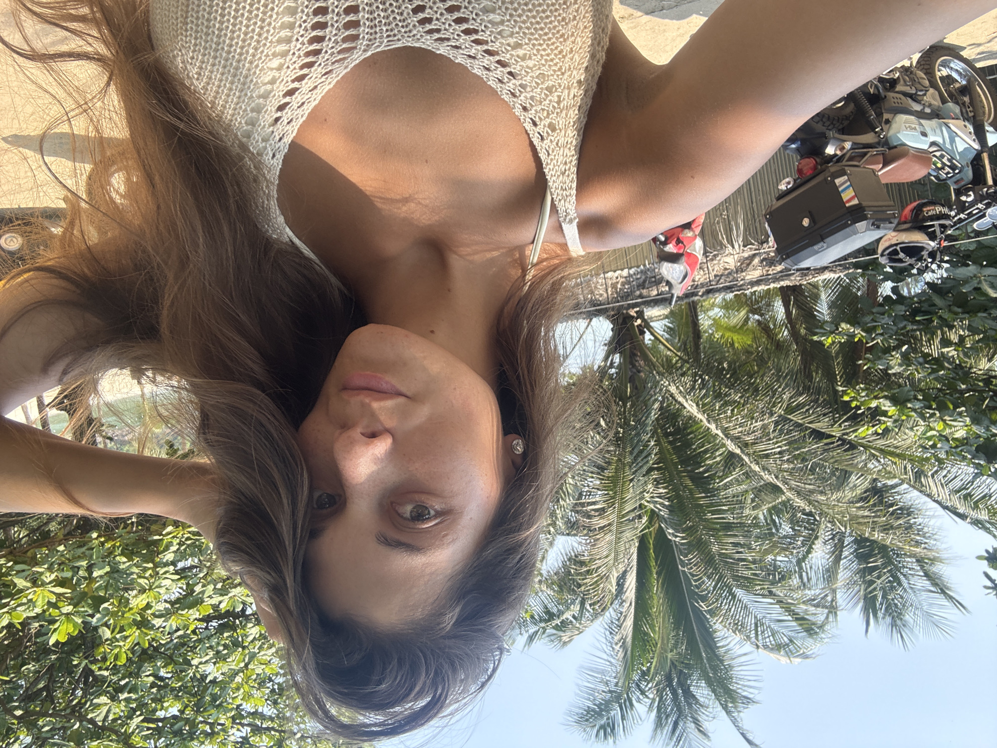 A selfie shows a woman in a white, crocheted top, with long brown hair. Her face is angled downwards towards the camera, with noticeable freckles. Behind her are palm trees and other greenery against a blue sky. To the right of the frame, a parked motorcycle is visible, along with a helmet labeled "CafePHO". Based on the helmet label and overall imagery, the location appears to be Đà Nẵng, Vietnam.