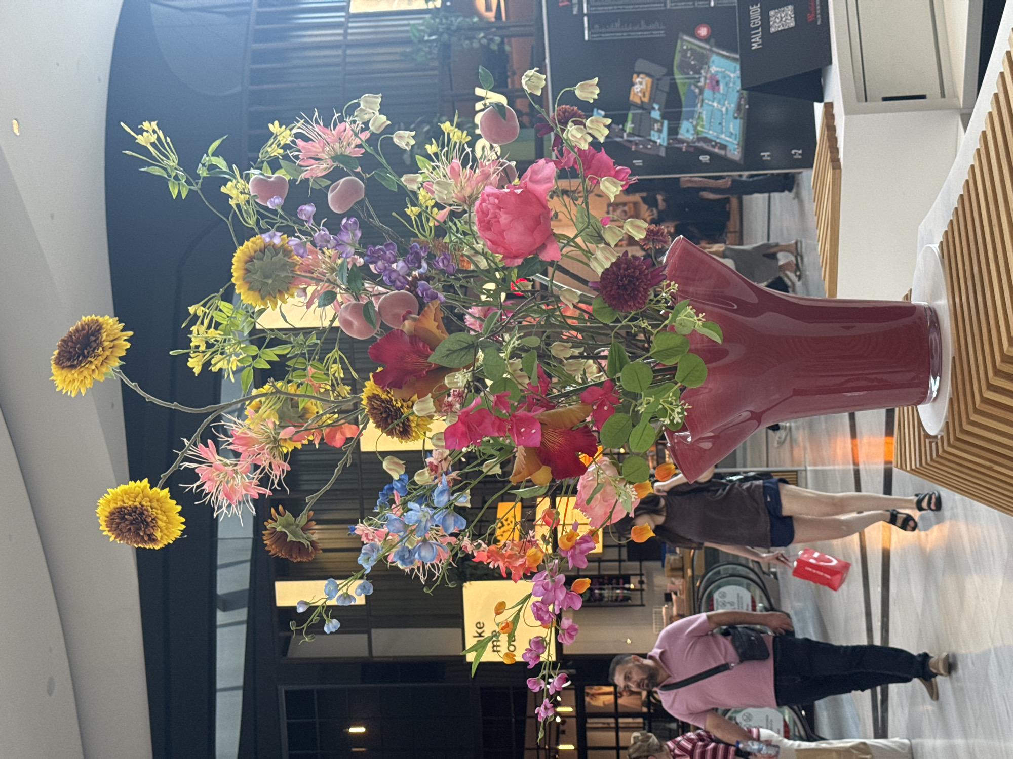 The image captures a vibrant floral arrangement in a large red vase, prominently displayed indoors. The flowers are a mix of colors including yellow, pink, purple, and blue, with some resembling sunflowers, peonies, and delphiniums. Small, peach-colored decorative fruits are interspersed among the blossoms. The background suggests an indoor public space, possibly a shopping mall, with architectural elements like a curved white wall and vertical wooden paneling.
In the background, several people are visible. One woman in a brown top and blue shorts is walking, holding a red shopping bag. Another person, wearing a pink polo shirt and dark pants, is walking towards the right side of the frame. A third person, wearing a striped shirt, is also visible in the lower part of the image. The lighting indicates it is daytime. A sign with "MALL GUIDE" and a map is also visible on the wall, further suggesting a mall setting. The overall impression is one of a well-decorated public space.