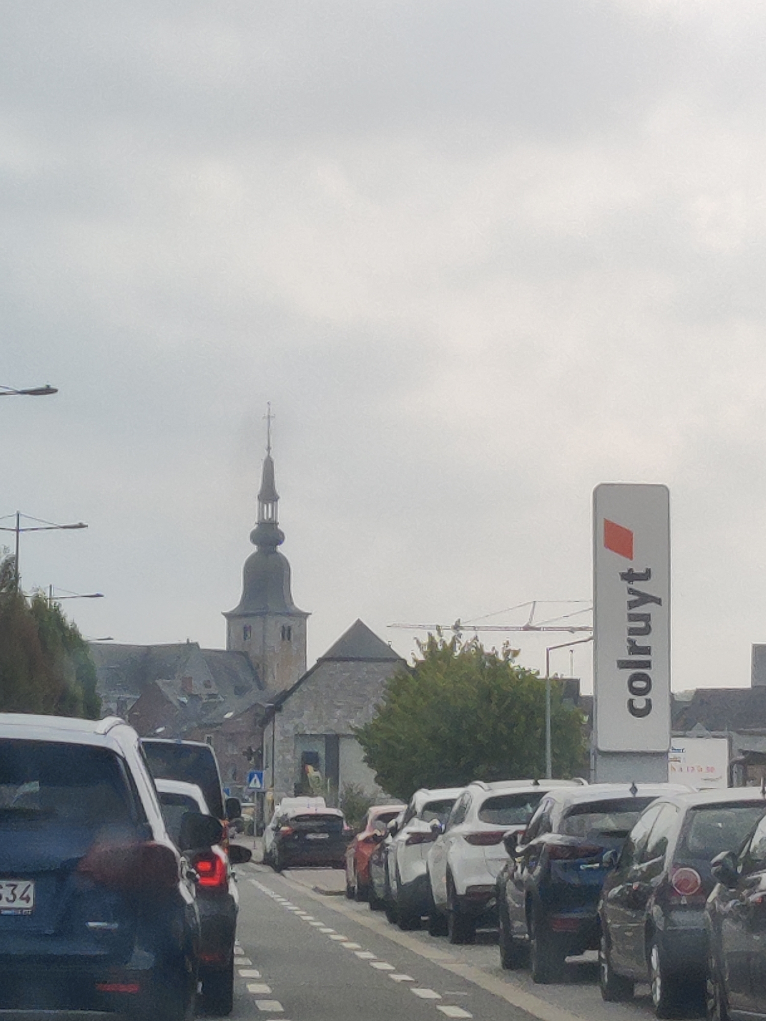 The image depicts a street scene in Marche-en-Famenne, Belgium, with a line of cars stopped in traffic. In the background, a church steeple rises above the buildings, and a sign for "colruyt" is visible on the right side of the frame. The sky is overcast, suggesting a cloudy day. The cars are diverse in make and model, and some have their brake lights illuminated, indicating they are stationary. The overall impression is one of a typical urban or semi-urban environment experiencing a traffic slowdown.