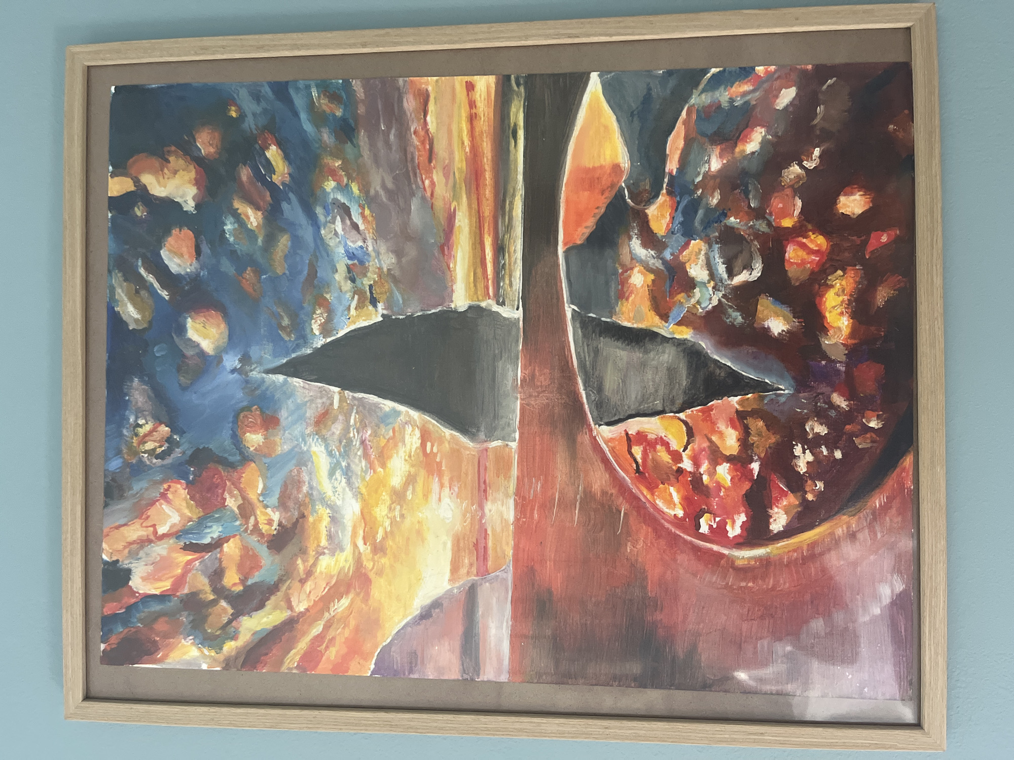 This is a framed abstract painting. The painting is divided vertically into two halves, with a central dark area that appears to be torn or ripped. The left half features a blend of dark blues, with bright orange and yellow splotches, possibly representing light or fire. The right half is dominated by deep reds and browns, with similar bright orange and yellow accents. There is a distinct curved shape on the right side, resembling a bowl or a vessel. The overall impression is one of abstract forms and vibrant colors. The painting is housed in a light-colored wooden frame and is displayed against a plain blue wall. There is no visible text.