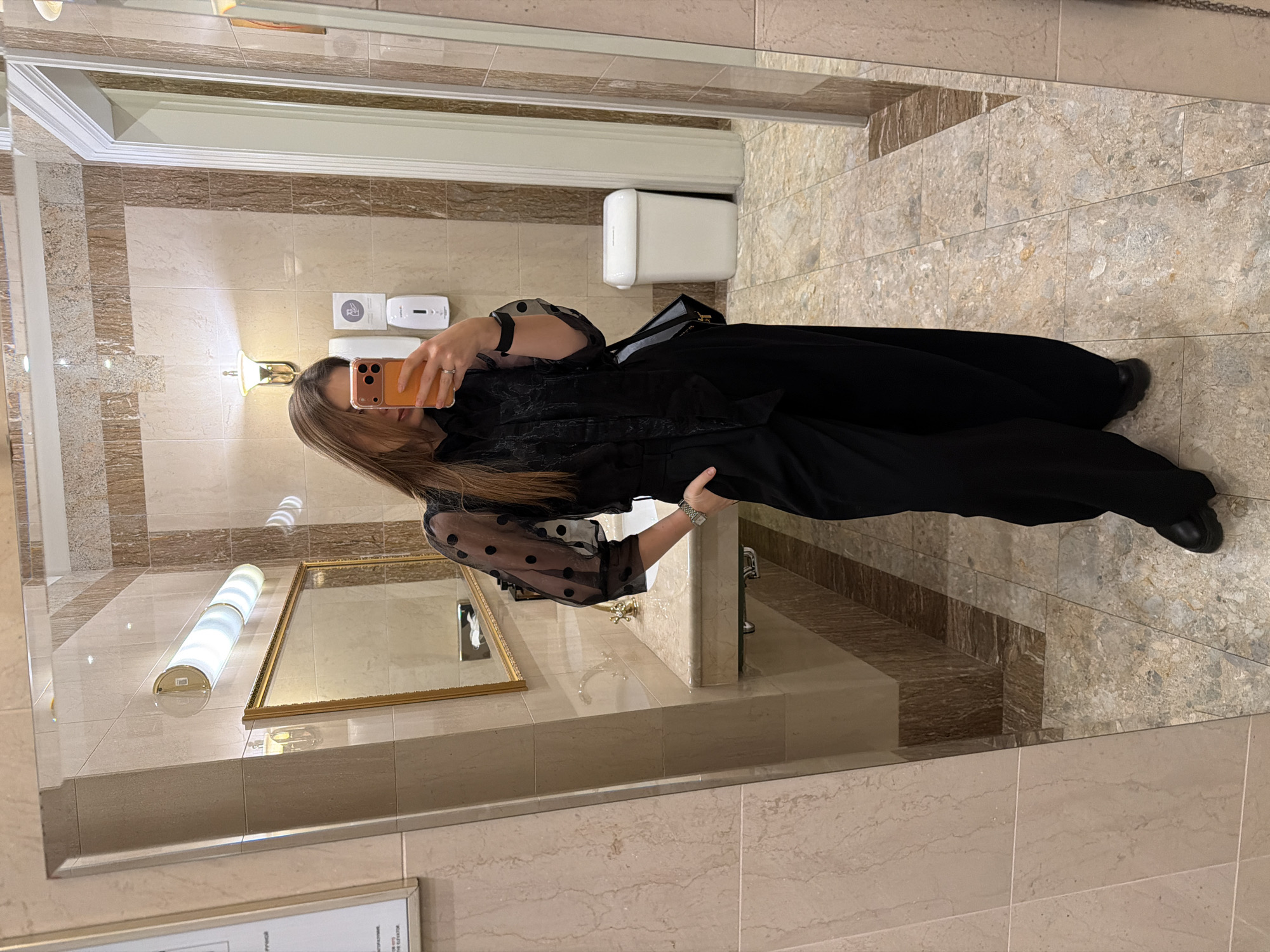 A full-length reflection of a woman in a bathroom, located in an unknown city. The woman holds an orange phone in her right hand, presumably taking a photo. She is wearing a black top with polka dot sheer sleeves and long black pants. A black purse hangs on her right shoulder. The bathroom has beige tile floors and walls with brown striped accents. A gold-framed mirror hangs above the sink, which is made of a light-colored marble or stone. A soap dispenser and toilet paper dispenser are mounted on the wall. A light fixture is visible above the mirror.