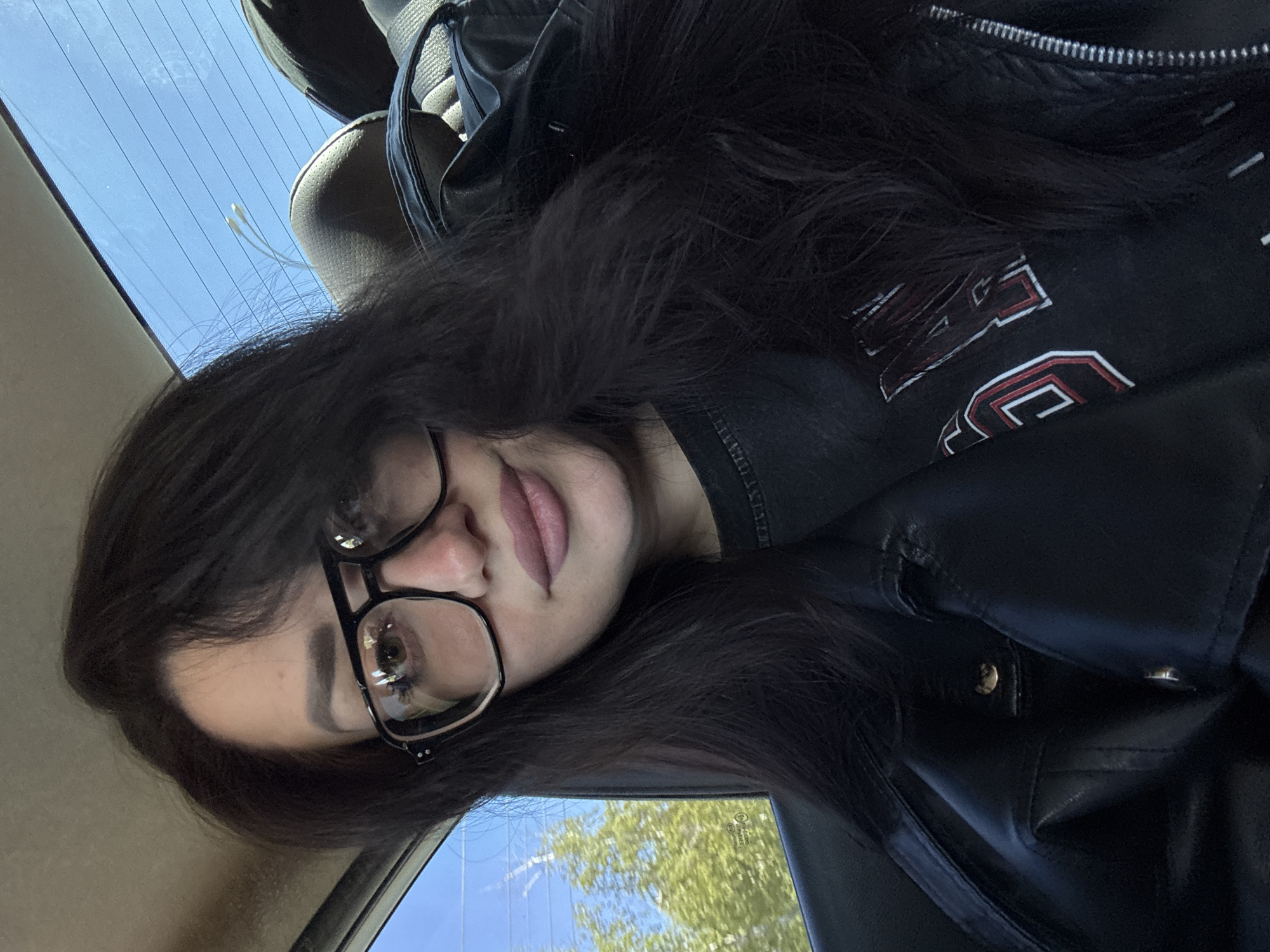A person is sitting in a car, with their head tilted to the side. They are wearing glasses and have dark hair. The person is wearing a black t-shirt and a black leather jacket. The car window is visible in the background, with a view of trees and a blue sky. The location is unknown.