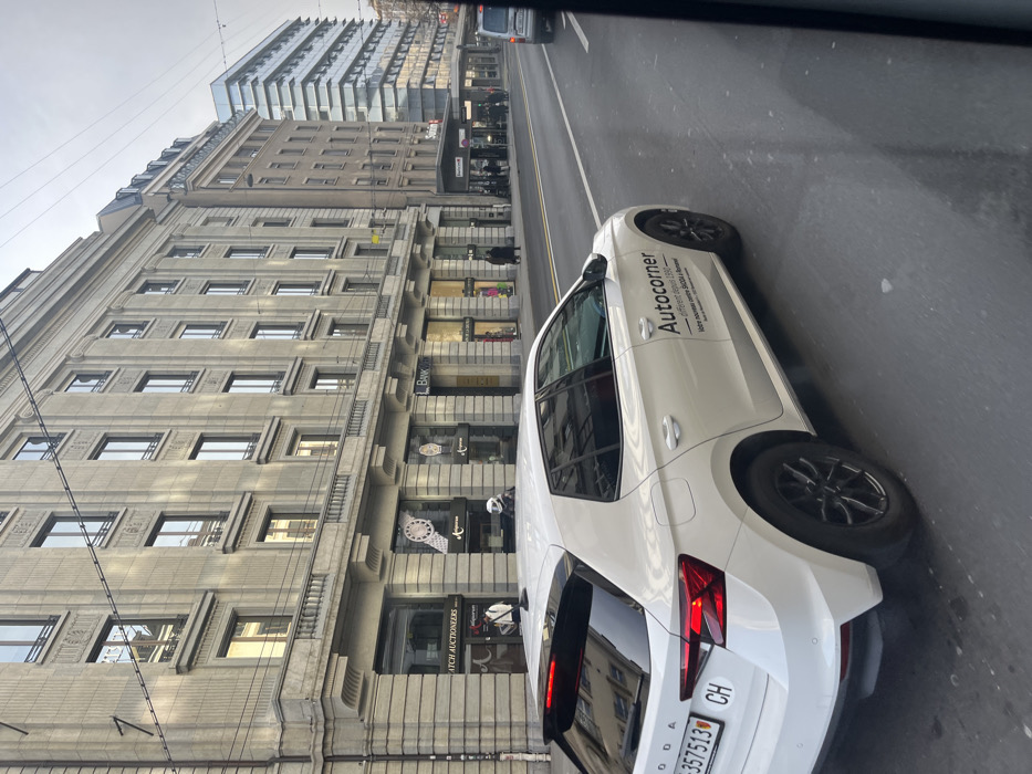The image captures a daytime street scene in an urban environment, likely in Geneva, Switzerland, viewed from inside a moving or parked vehicle.
In the foreground, a white, modern-looking hatchback car, identified as a Skoda by the logo on its trunk, occupies the right side of the frame. The car has "Autocorner" written in black text on its side, along with smaller, unreadable text below it. The Swiss license plate "CH VD 357513" is clearly visible on the rear. The car features black multi-spoke wheels.
To the left of the street, a tall, classical-style building with many floors and numerous rectangular windows dominates the mid-ground. Its facade appears to be made of light-colored stone or concrete, adorned with architectural details. The ground floor of this building houses several retail storefronts, including one labeled "WATCH AUCTIONEERS" and another partially visible as "Bankor." Display windows showcase various items, including a large watch face. A few indistinct figures, likely pedestrians, can be seen on the sidewalk in front of these shops, one appearing to stand near a storefront or entering it. Another person on a small scooter or motorcycle is visible near the sidewalk.
Further down the street, behind the classical building, a more contemporary building with a distinctive glass and metal facade is visible, reflecting the overcast sky. The road surface is asphalt, appearing slightly damp in places. Overhead power lines or tram wires stretch across the top portion of the frame. The sky is uniformly gray and overcast, suggesting a cloudy day with diffused light. The overall scene depicts typical city life with traffic and commercial activity.