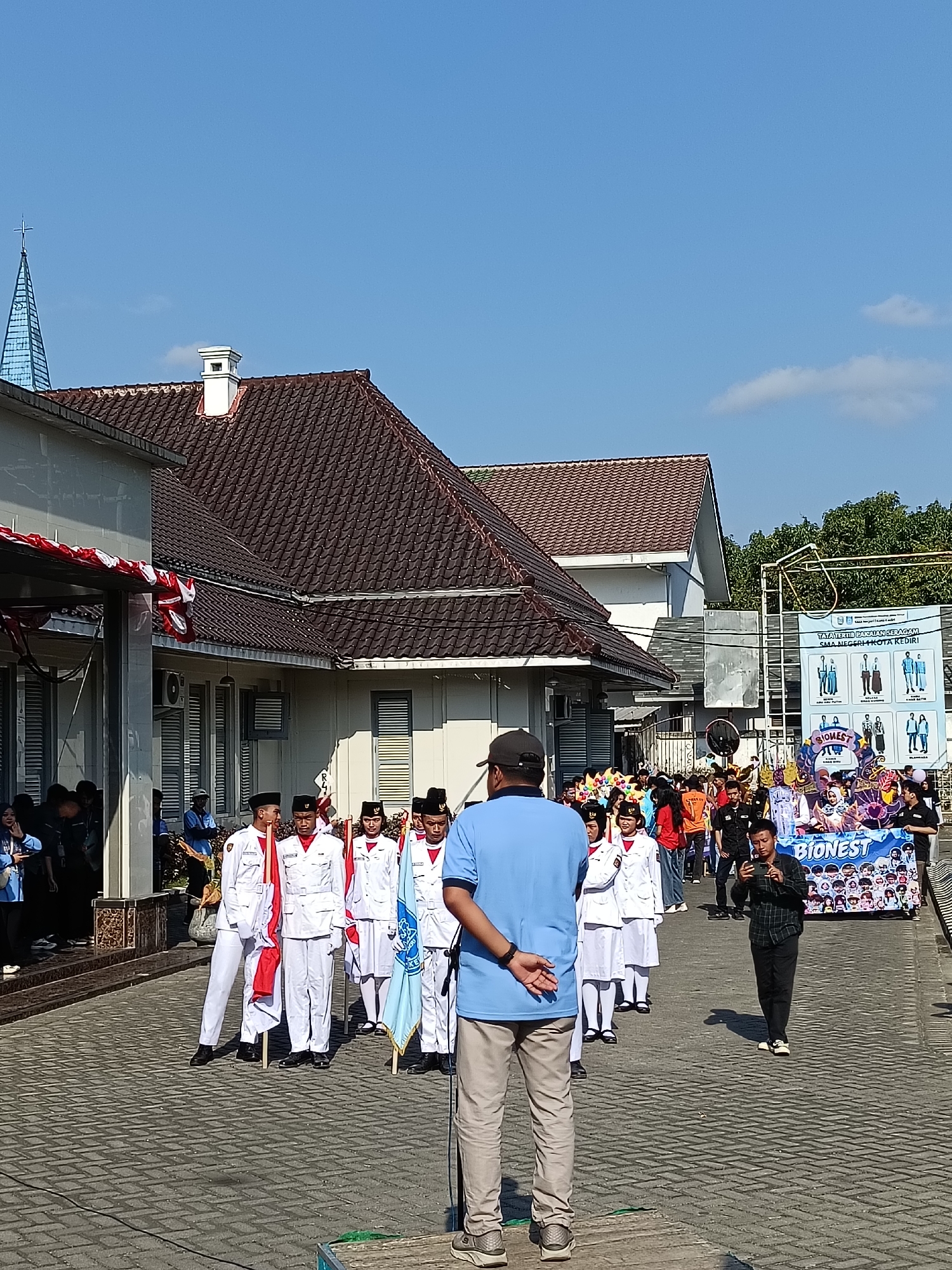 A group of students in white uniforms are participating in a parade in front of a building with a brown tiled roof and a white chimney. The students are holding flags and appear to be marching. In the foreground, a man wearing a blue shirt and tan pants is standing with his back to the camera, facing the parade. A banner in the background displays information about "BIONEST" and lists different school uniforms, suggesting the parade is related to a school event. The sky is clear and blue. The scene appears to be taking place in a public area, possibly a street or courtyard, paved with interlocking bricks. The architectural style of the building and the presence of the banner indicate this is likely a location in Kediri City, Indonesia.
