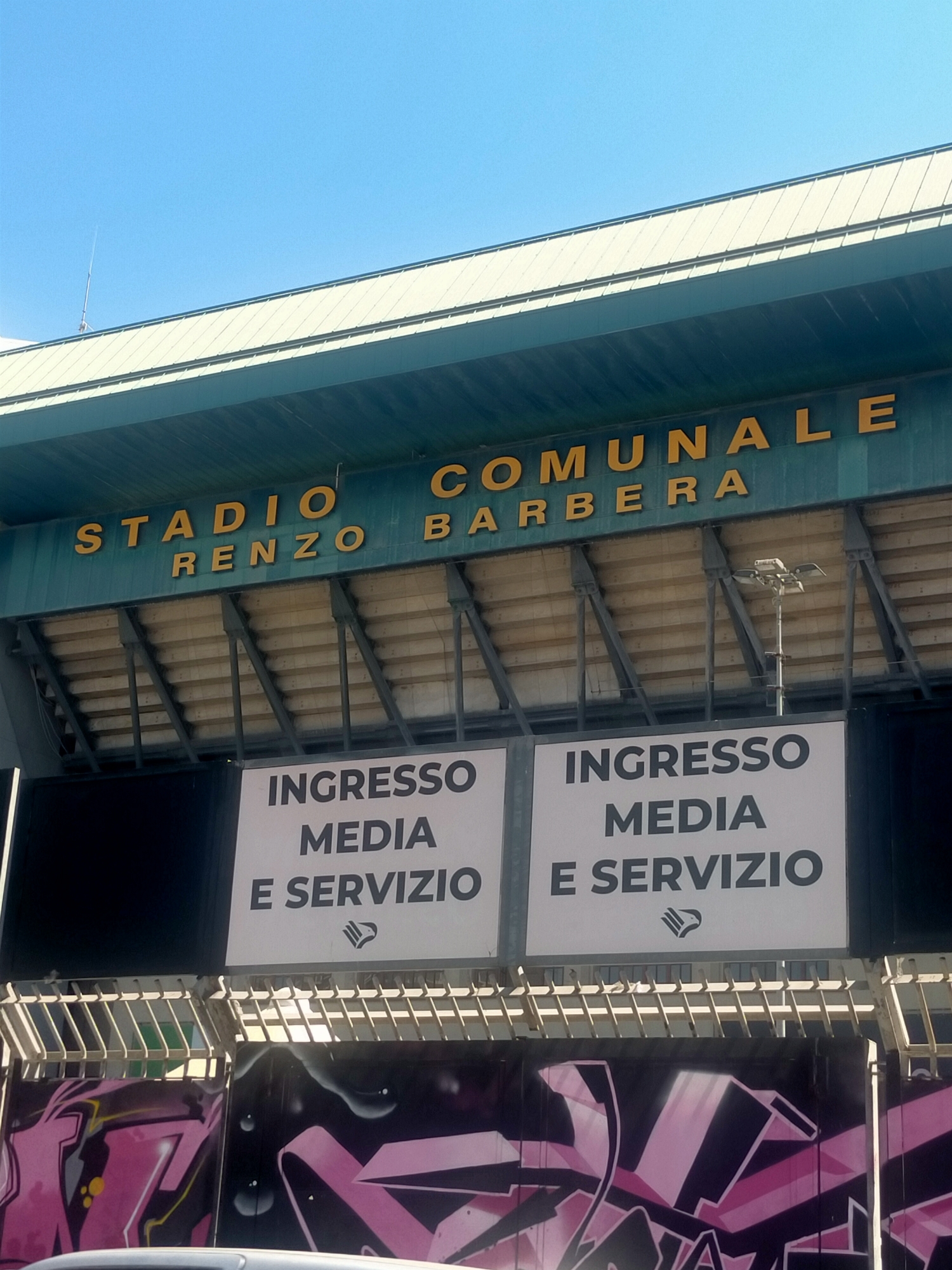 The image shows the exterior of the Stadio Renzo Barbera in Palermo, Italy, under a clear blue sky. The stadium's green facade features large yellow lettering that reads "STADIO RENZO BARBERA COMUNALE". Below this sign, two large white panels are visible, each with black text. The left panel reads "INGRESSO MEDIA E SERVIZIO", and the right panel also reads "INGRESSO MEDIA E SERVIZIO". A small logo, resembling a bird in flight, is present below the text on the right panel. The stadium seating is visible in the background behind the signs, supported by metal beams. At the very bottom of the image, a section of a graffiti mural with pink and black abstract designs can be seen. There are no people or animals depicted in the image. The overall environment is outdoors, suggesting a sunny day.